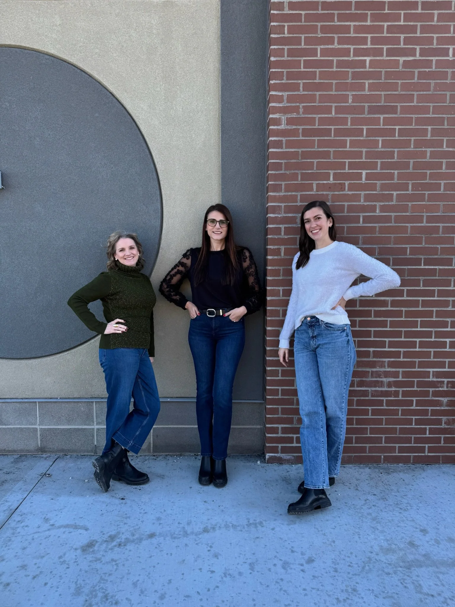 Three women standing together outdoors against a brick and concrete wall, smiling.
