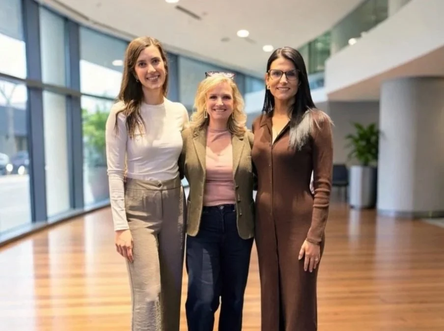 Three women standing together inside a modern building with large windows and wooden floors, smiling at the camera.