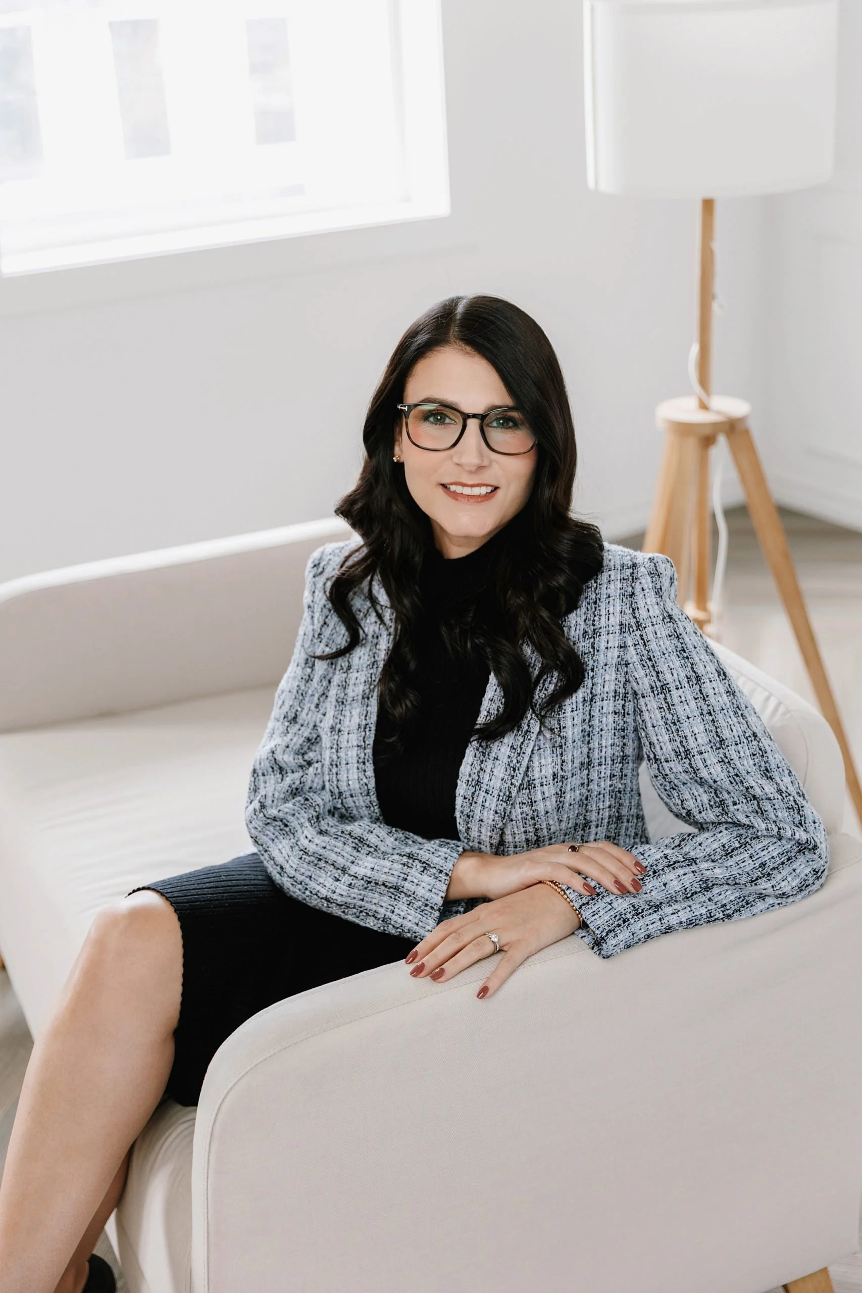 Portrait of a woman with dark hair, glasses, and a blazer, sitting on a cream-colored sofa in a bright, modern room with large windows and a standing lamp.