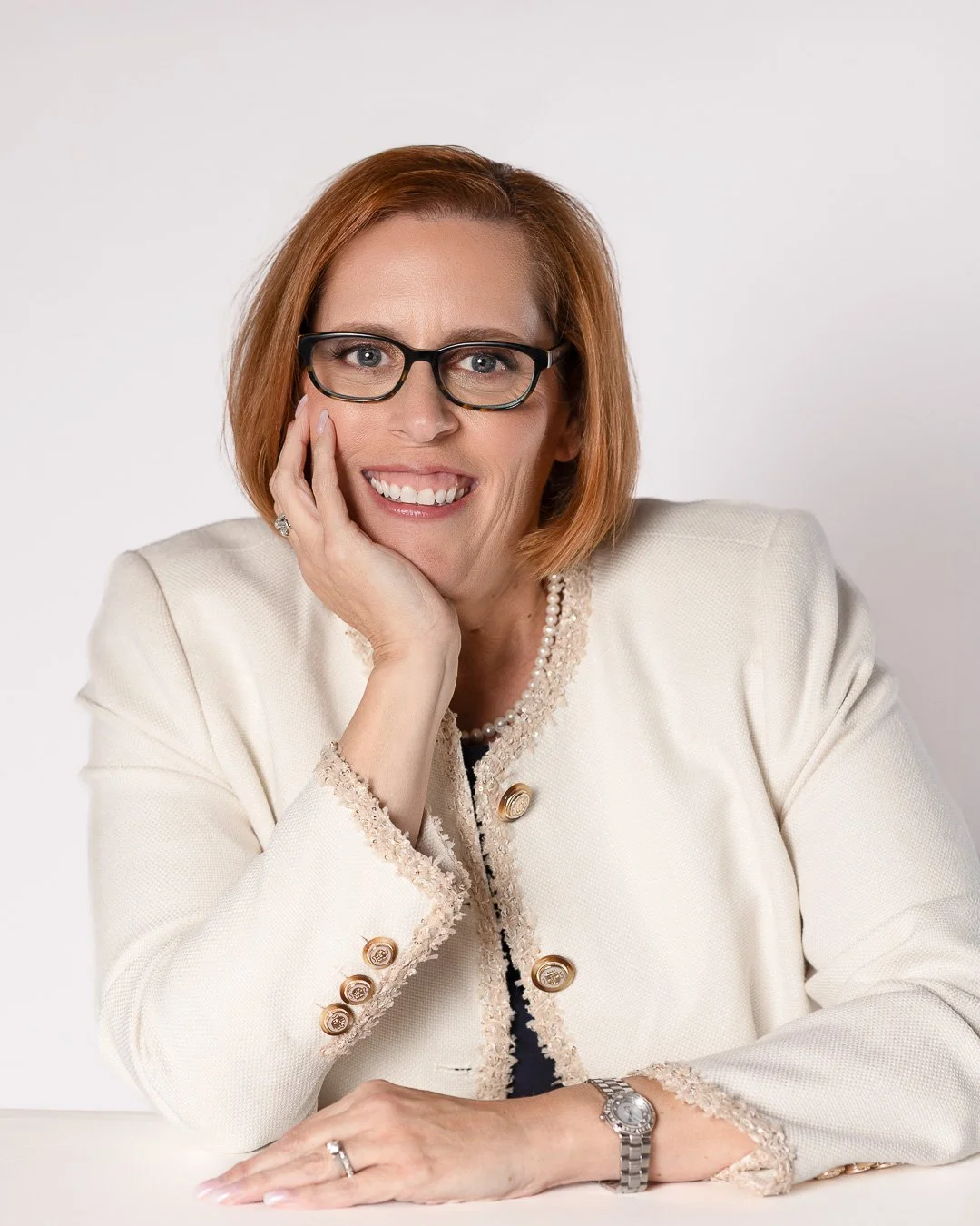 A woman with red hair and glasses smiling, resting her chin on her hand, wearing a cream-colored jacket with decorative buttons and a pearl necklace, against a plain white background.