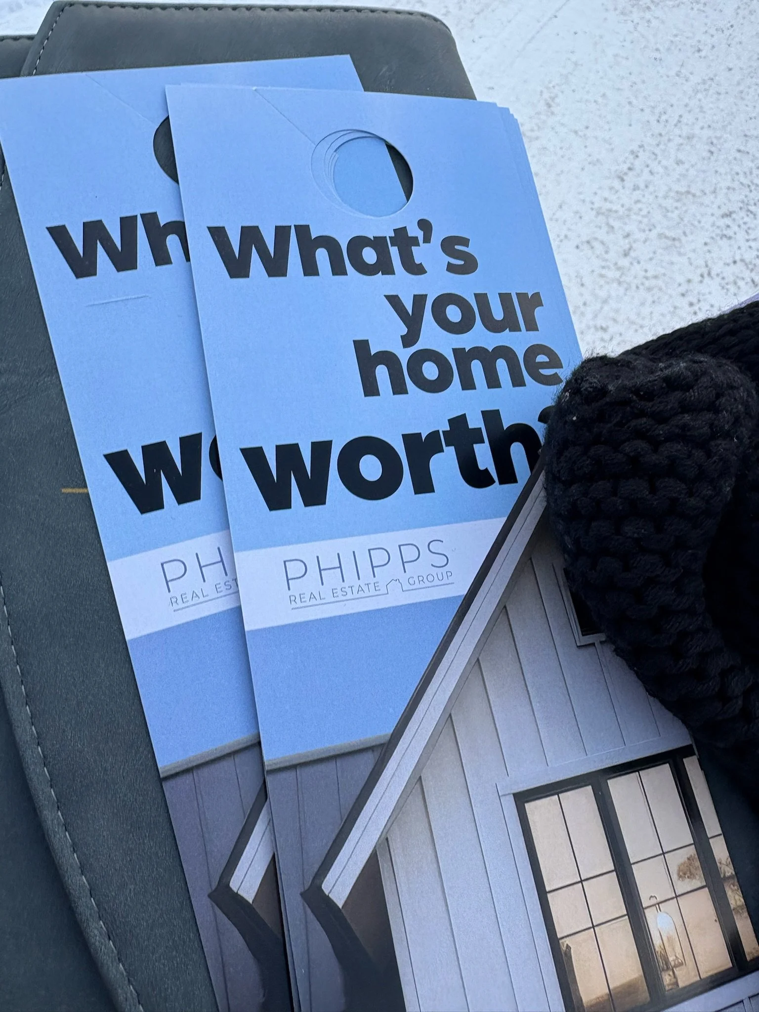 Two flyers for PHIPPS Real Estate Group with the text "What's your home worth?" on a dark surface, next to a black knitted item and a brochure with an image of a house exterior.