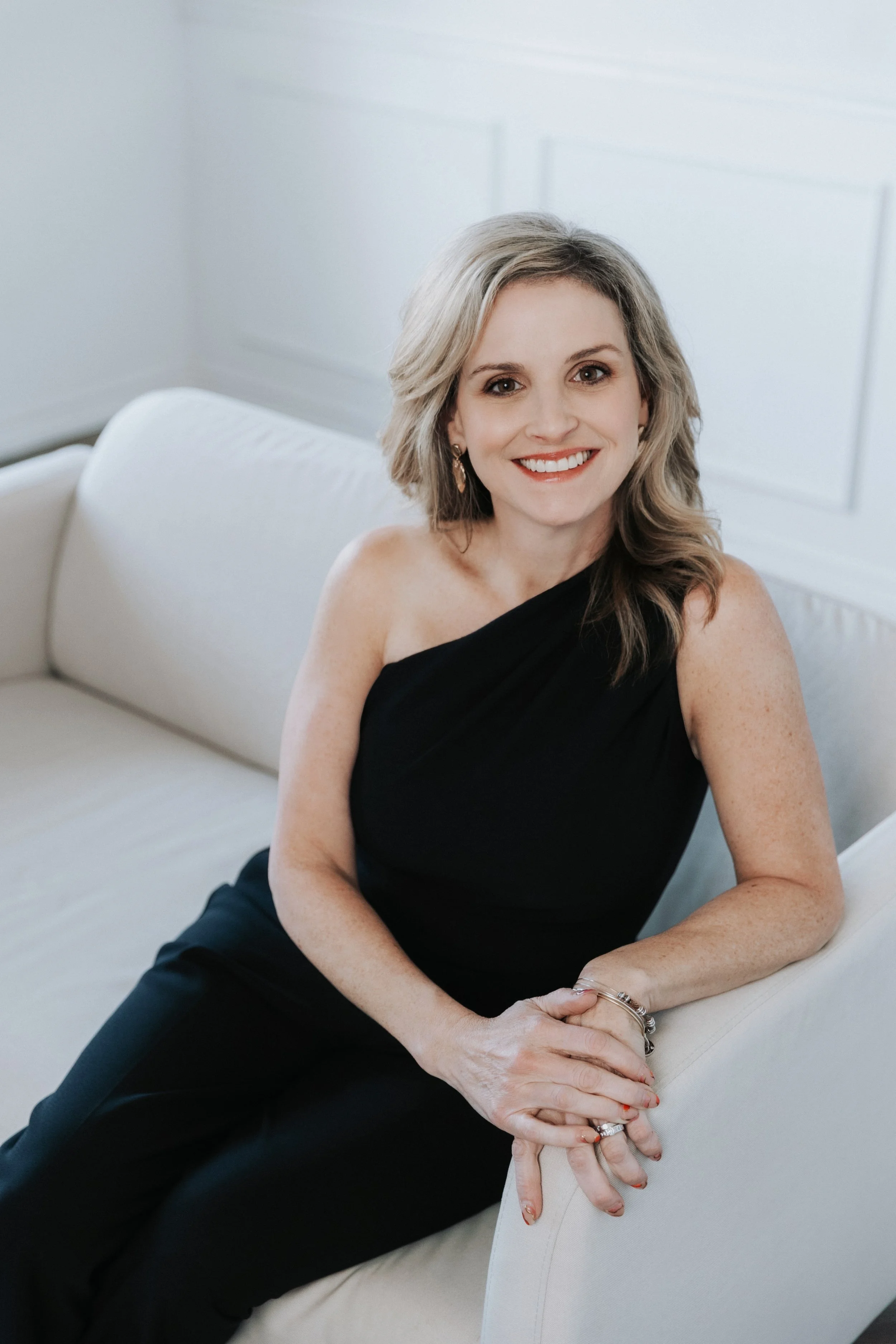 A woman in a black sleeveless dress sitting on a cream sofa, smiling at the camera in a bright white room.