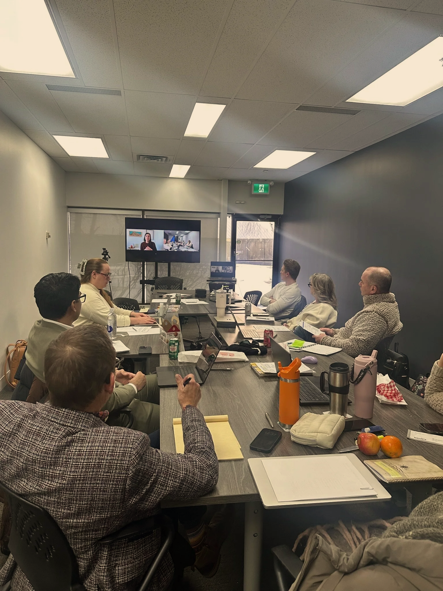 People attending a virtual meeting in a conference room with laptops, notebooks, and drinks on the table.
