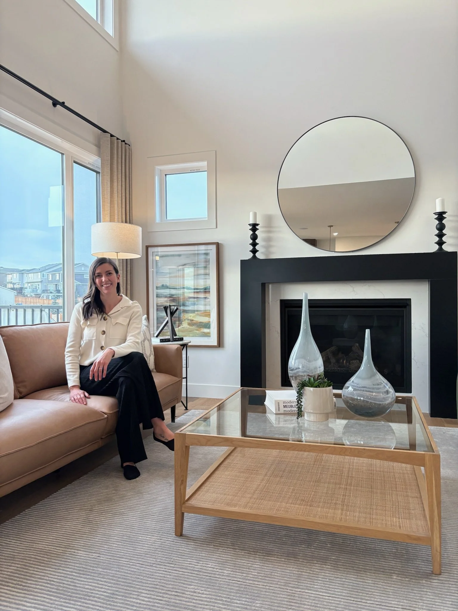 A woman sitting on a beige sofa in a modern living room with a glass coffee table, fireplace, round mirror, and large windows showing a neighborhood view.