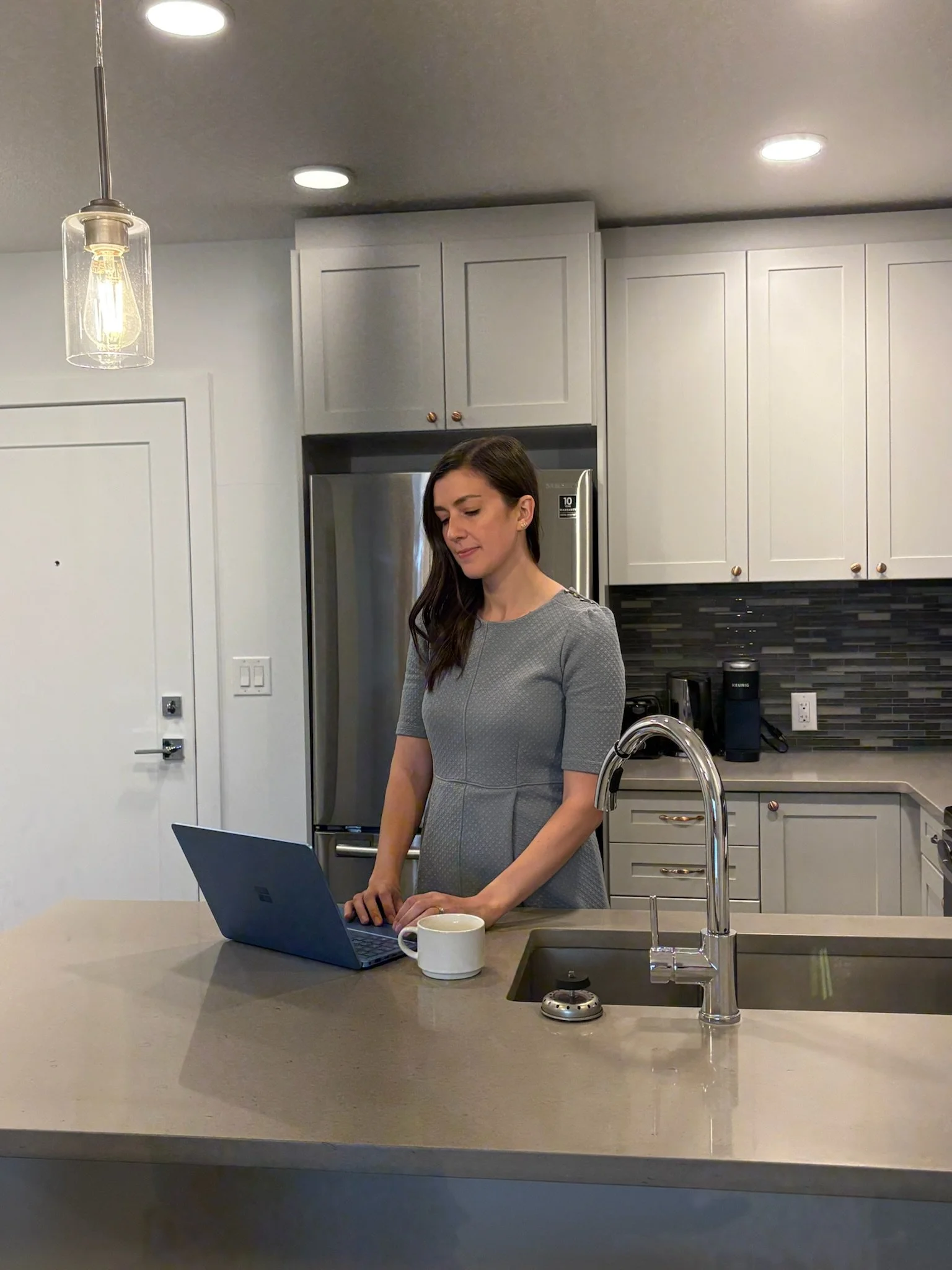 A woman in a gray dress standing at a kitchen island with a laptop and a coffee mug, kitchen with gray cabinets, a stainless steel refrigerator, and a black kitchen appliance in the background.