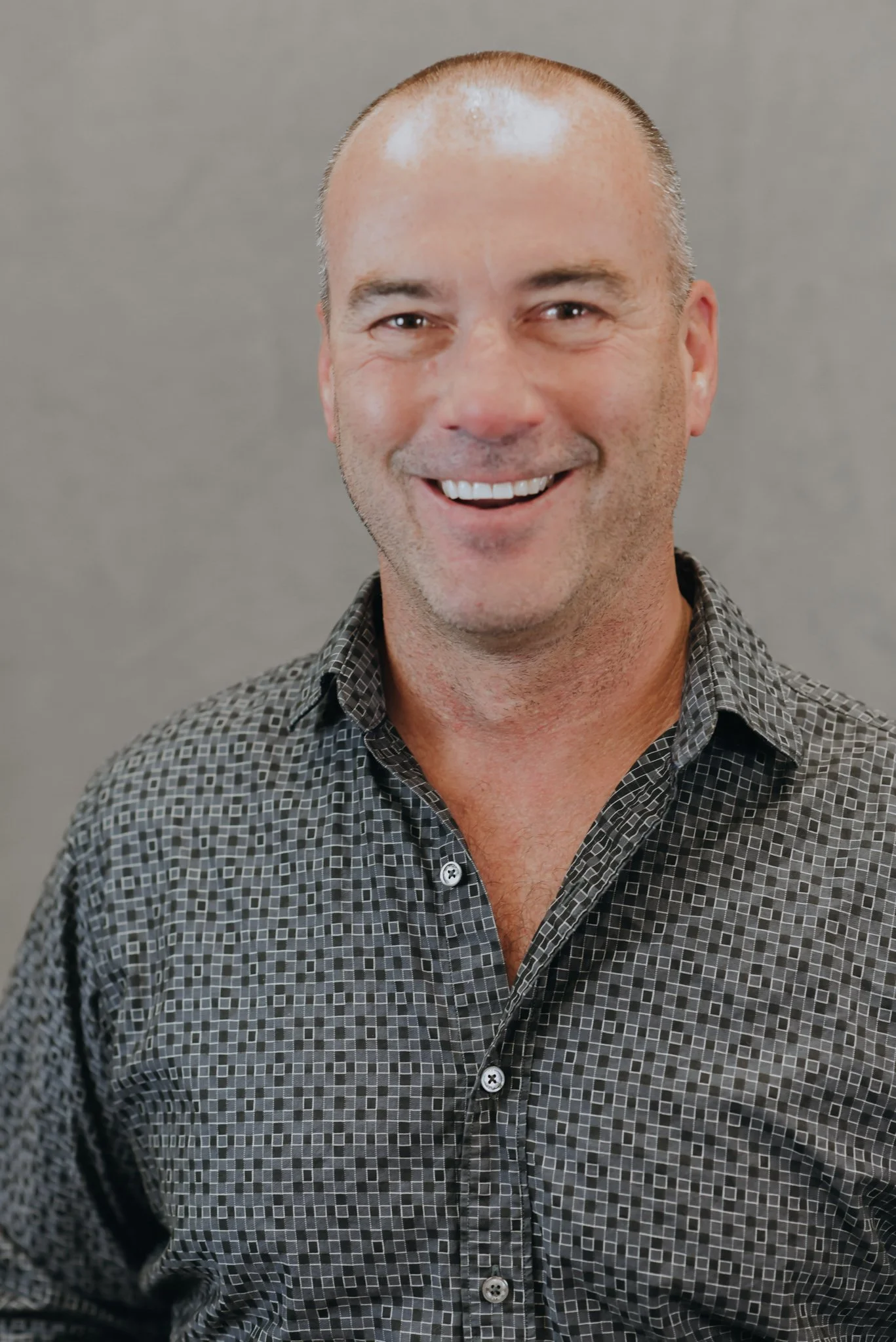 A smiling man with a shaved head, wearing a checkered button-up shirt, standing against a plain gray background.