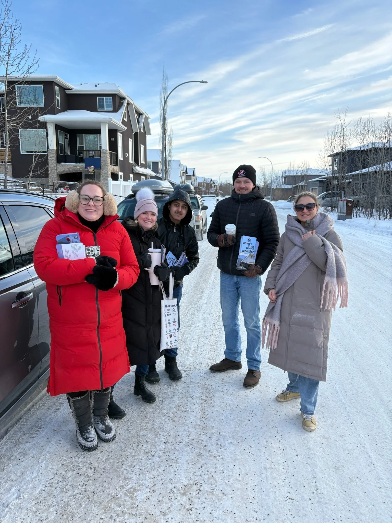 Group of five people standing on a snowy street in winter, smiling and holding flyers and cups, with residential houses in the background.