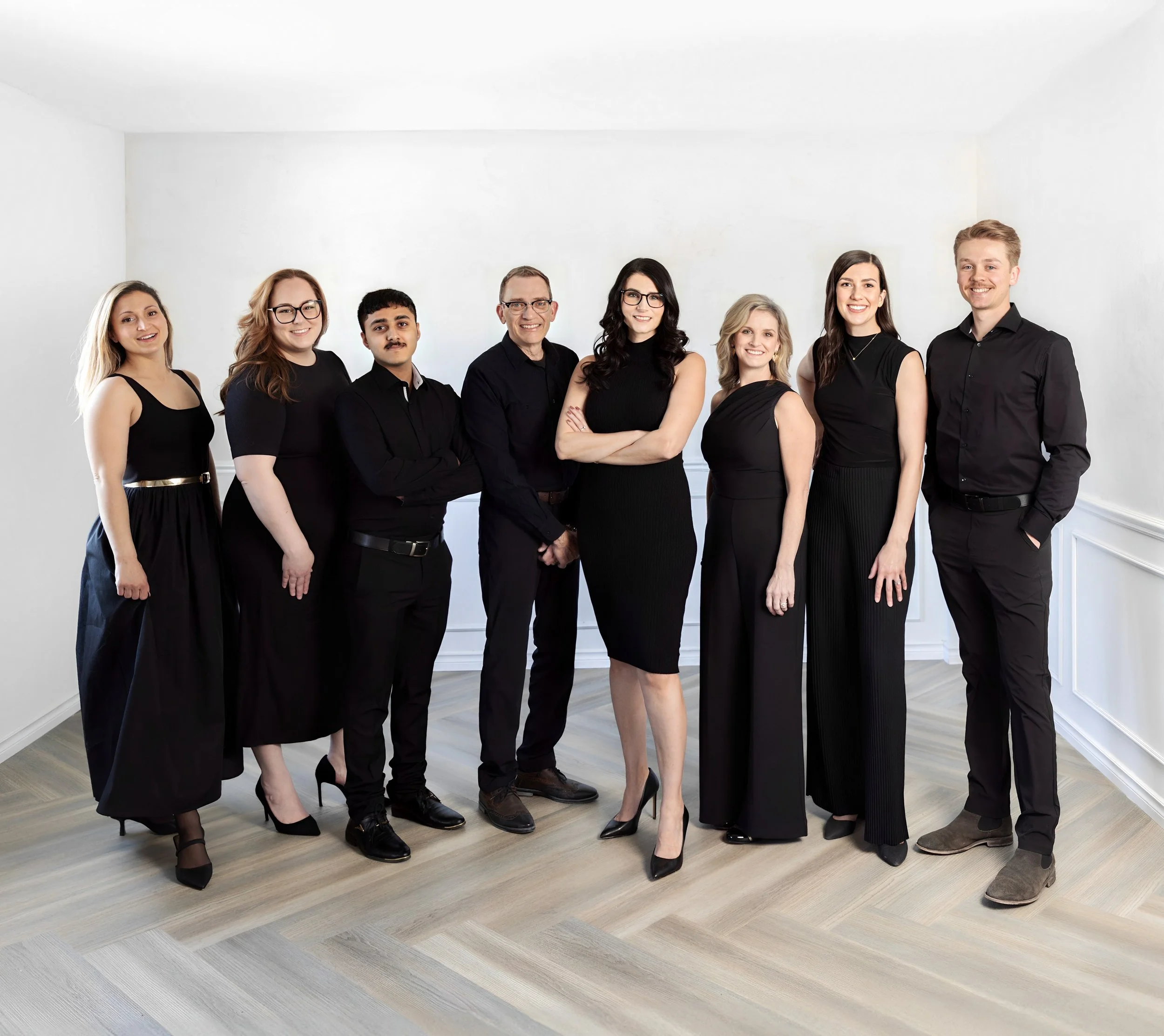 Group of nine diverse people dressed in black, standing together in a white room, smiling at the camera.