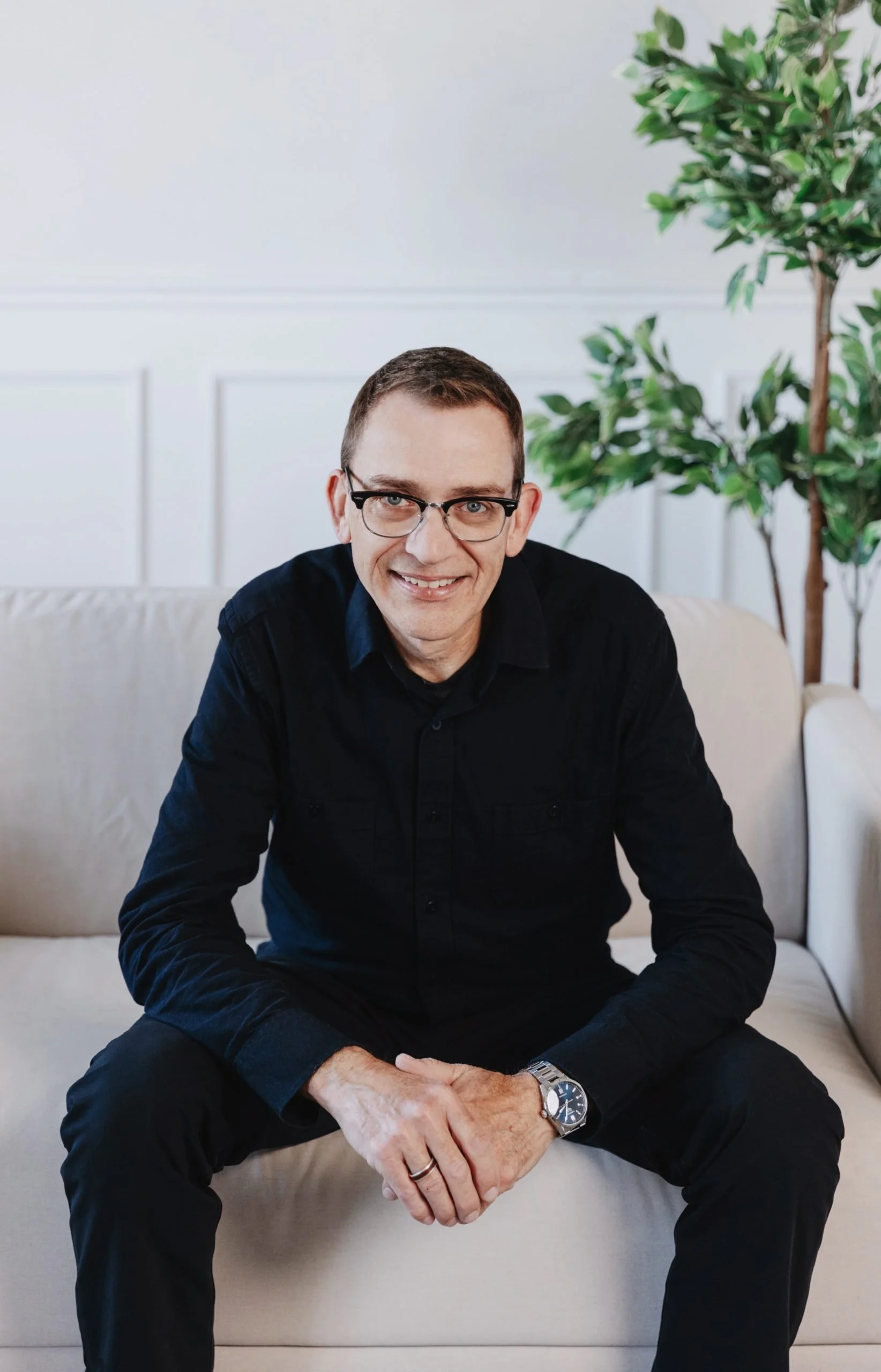 A man with glasses sitting on a beige sofa, smiling, with a plant in the background.