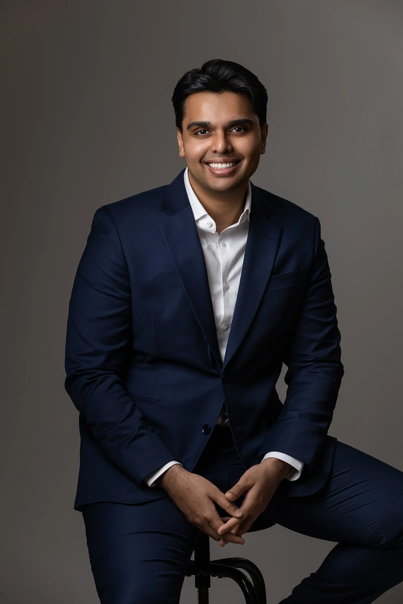 A man dressed in a navy blue suit sitting on a black stool against a gray background, smiling at the camera.
