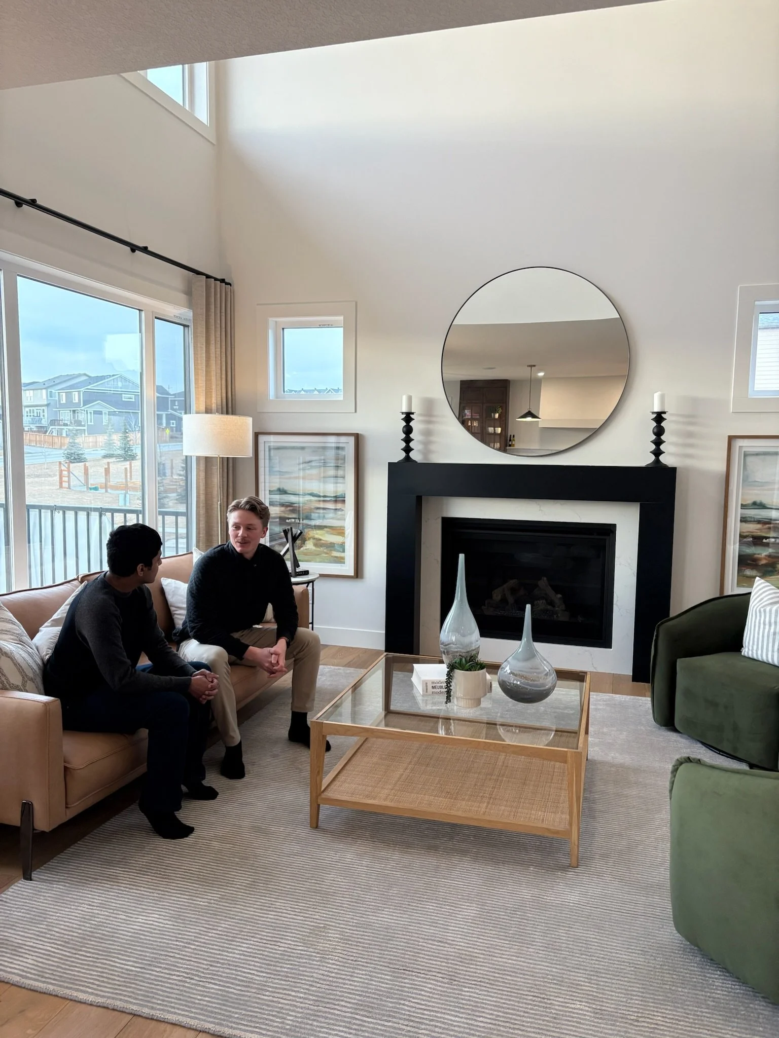 Two men sitting on a beige sofa in a modern living room, engaged in conversation. The room features large windows, artwork, a black fireplace with a round mirror above it, and a glass coffee table with decorative vases.