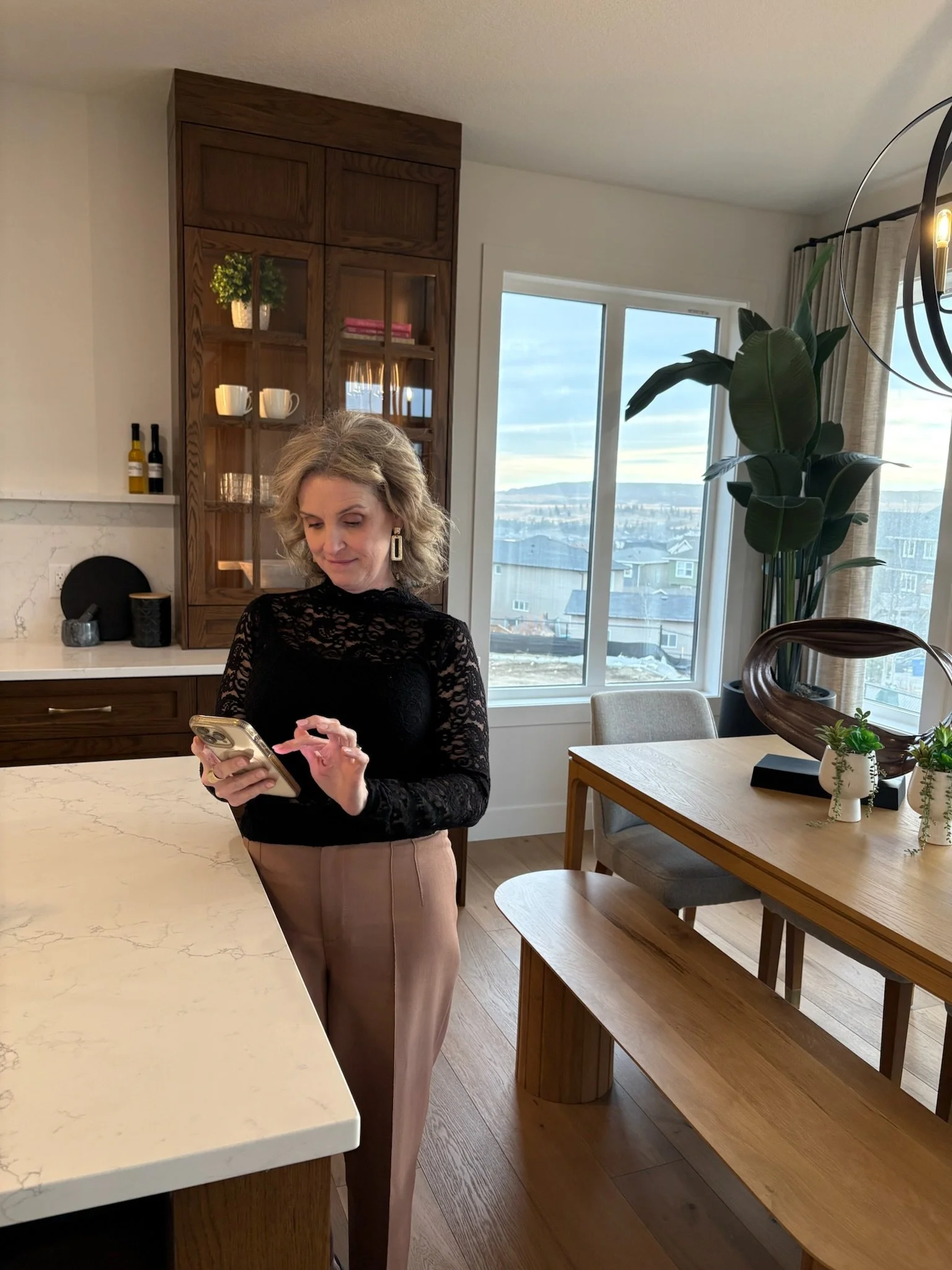 A woman with wavy blonde hair wearing a black lace long-sleeve top and beige pants standing in a modern kitchen, looking at her smartphone.