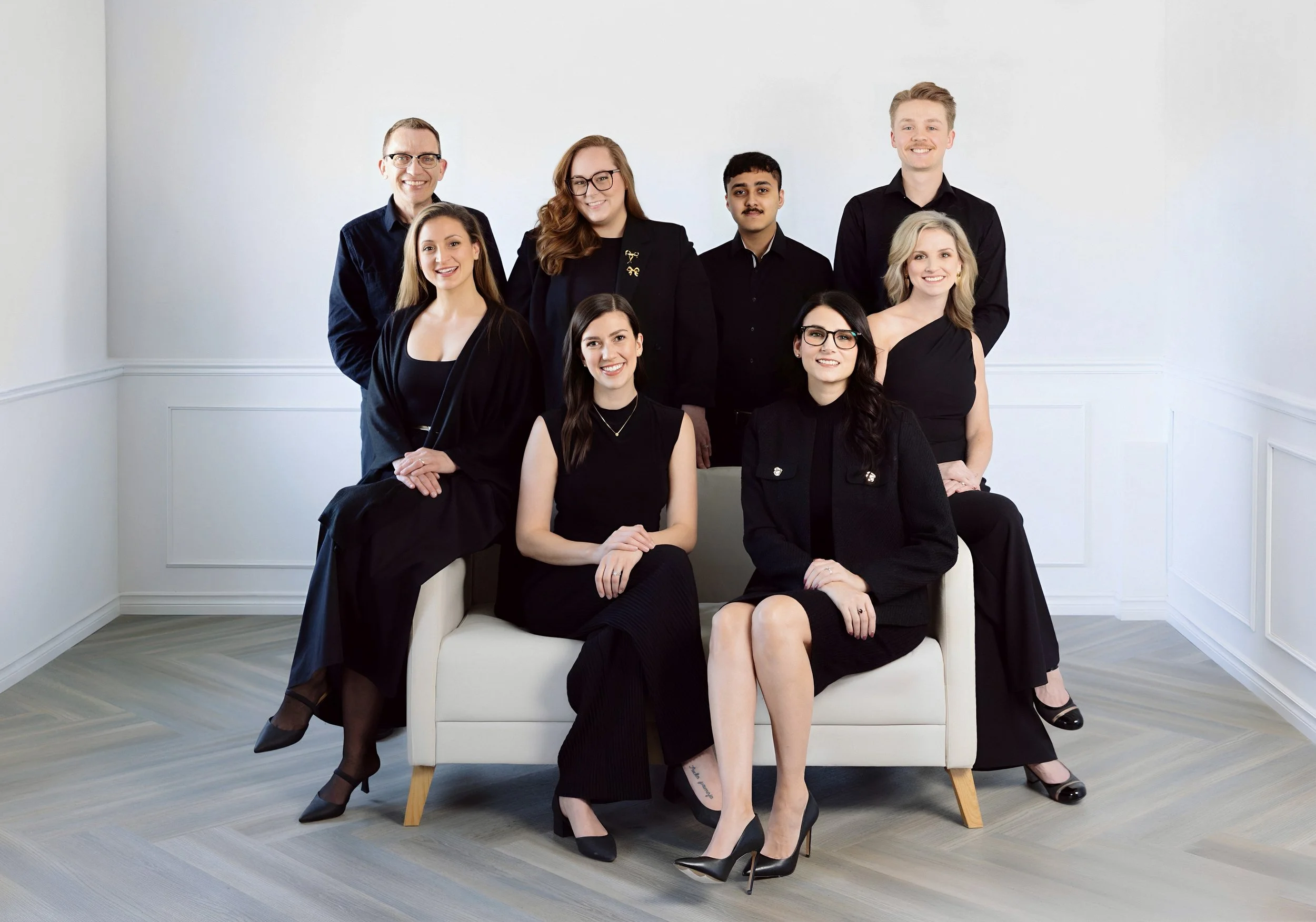 Group of nine professionally dressed people posing in a bright, minimalistic room with white walls and light wood floors.