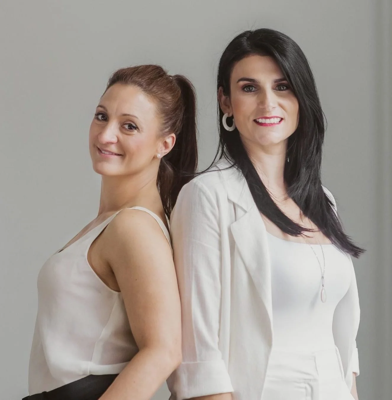 Two women standing back-to-back, smiling at the camera against a plain wall. One has brown hair in a ponytail and is wearing a sleeveless white top. The other has black hair, wears white earrings and a white blazer over a white top.