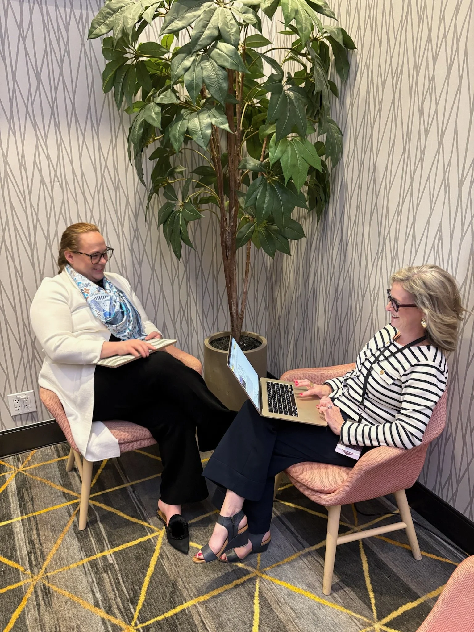Two women sitting in armchairs facing each other, engaged in conversation. One woman is holding a notebook, the other is using a laptop. They are in a room with patterned wallpaper, a large potted plant, and a carpet with a geometric design.