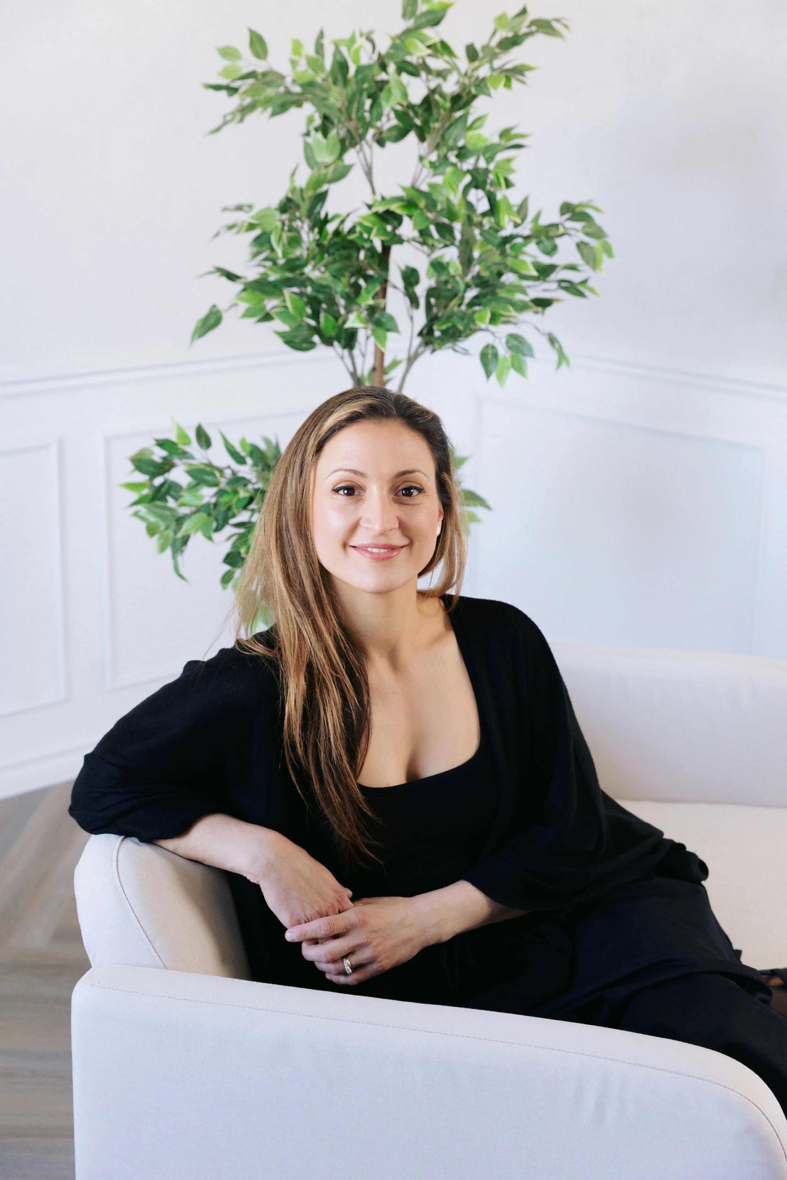 A woman with long brown hair, wearing a black top, sitting on a white sofa with her arm resting on the armrest, smiling at the camera. Behind her is a large green leafy plant in a white room.