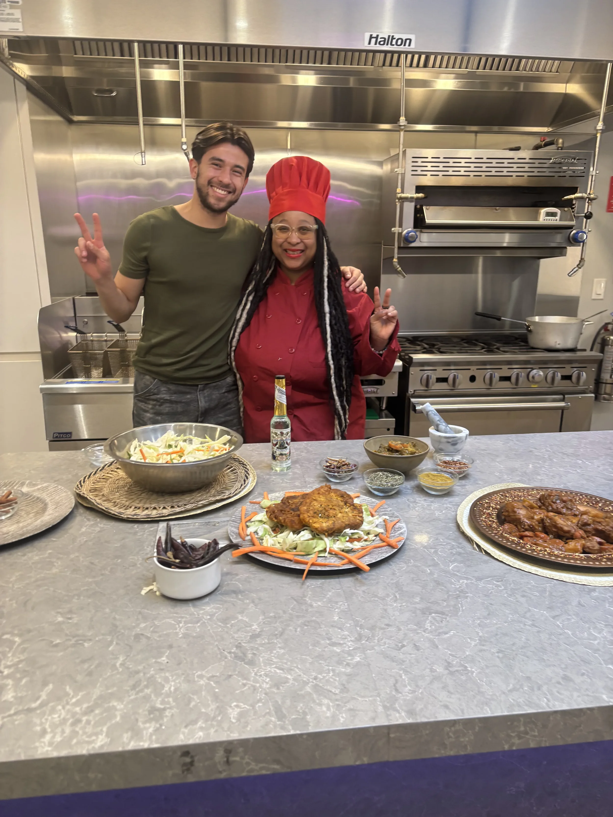 Two people, one man and one woman dressed as a chef, smiling and making peace signs in a commercial kitchen with food dishes on a marble countertop in front.
