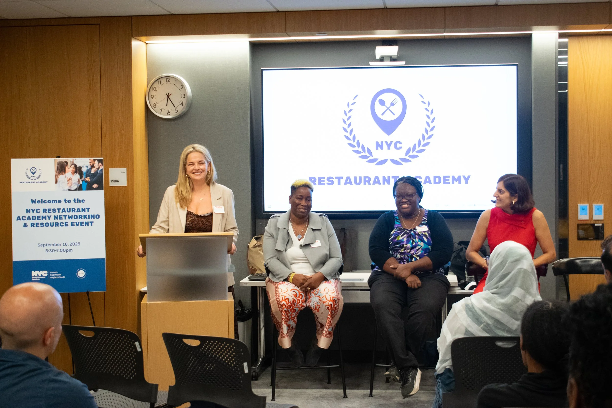 A woman stands at a podium smiling during a conference, with three women seated on stage behind her. The background displays a large screen with the NYC Restaurant Academy logo, showing a fork and spoon inside a location pin with laurel branches. To the left, a sign reads, 'Welcome to the NYC Restaurant Academy Networking & Resource Event, September 16, 2025, 5:30-7:00 pm.' The audience faces the stage, and the room has wood-paneled walls and a clock showing 6:15.