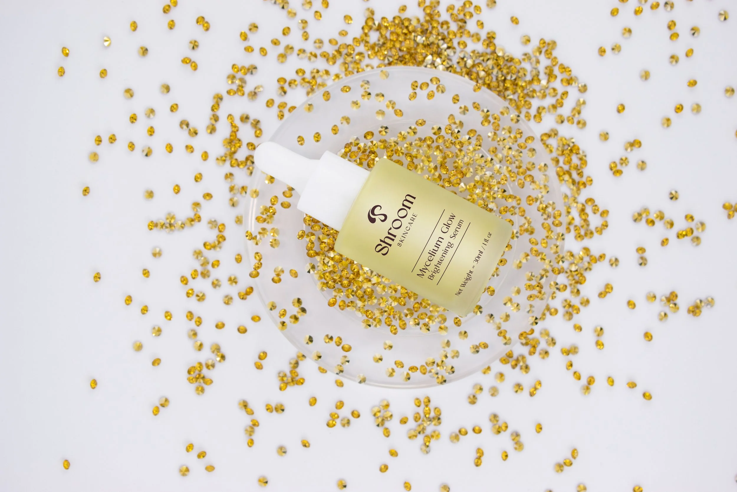A bottle of skincare serum labeled "ShroOm Skin Care Mycellum Glow Brightening Serum" is placed on a clear dish, surrounded by gold rhinestones scattered on a white surface.