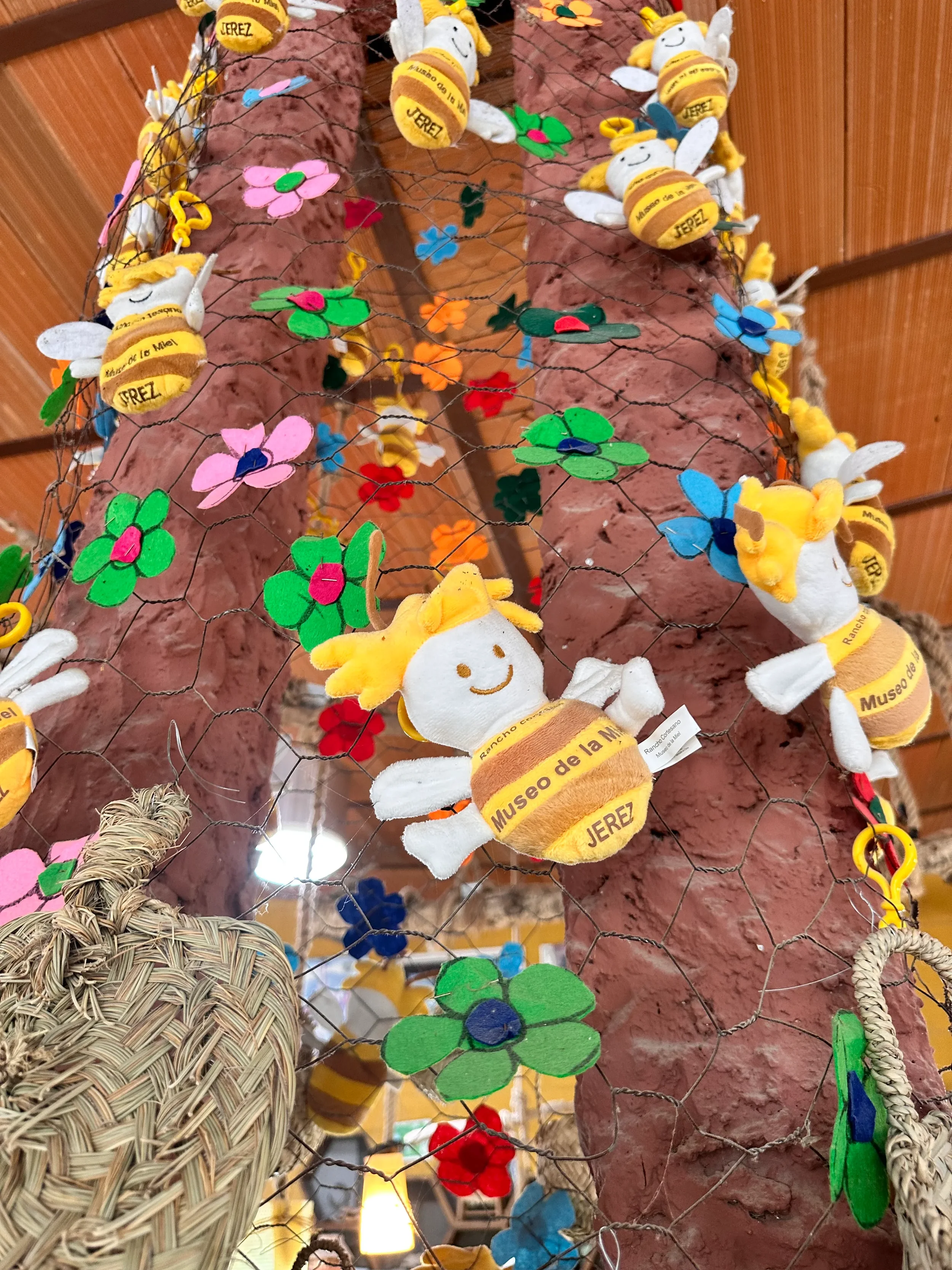 Colorful decorative items on a wire mesh, including plush bees with yellow and white bodies, smiles, and labels that say 'Museo de la Miel' and 'Jerez'; felt flowers in pink, green, blue, red, and orange; a woven basket; and a terracotta-colored background.