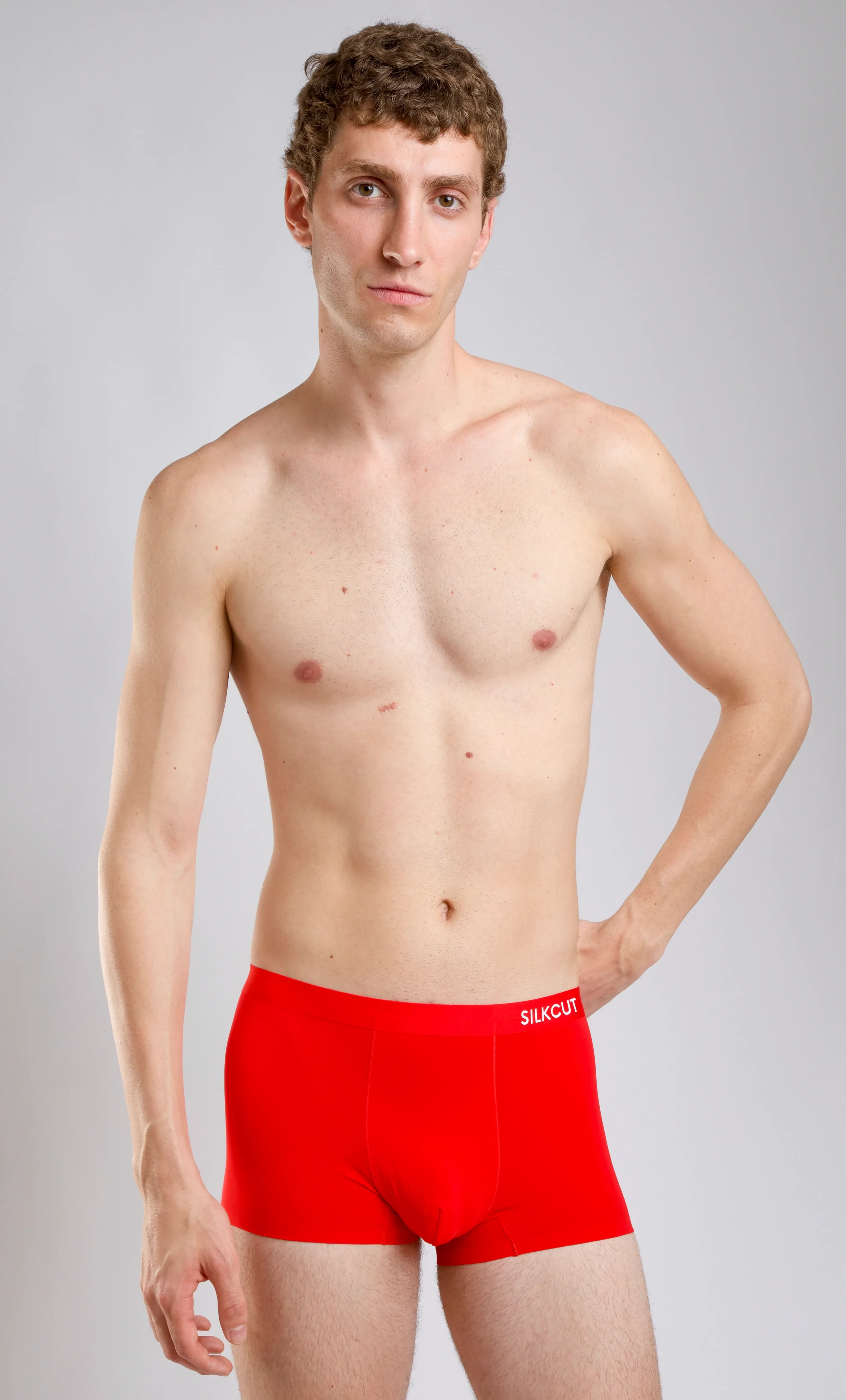 Studio fashion portrait of a male model standing in red Silkcut boxer briefs under clean indoor lighting.