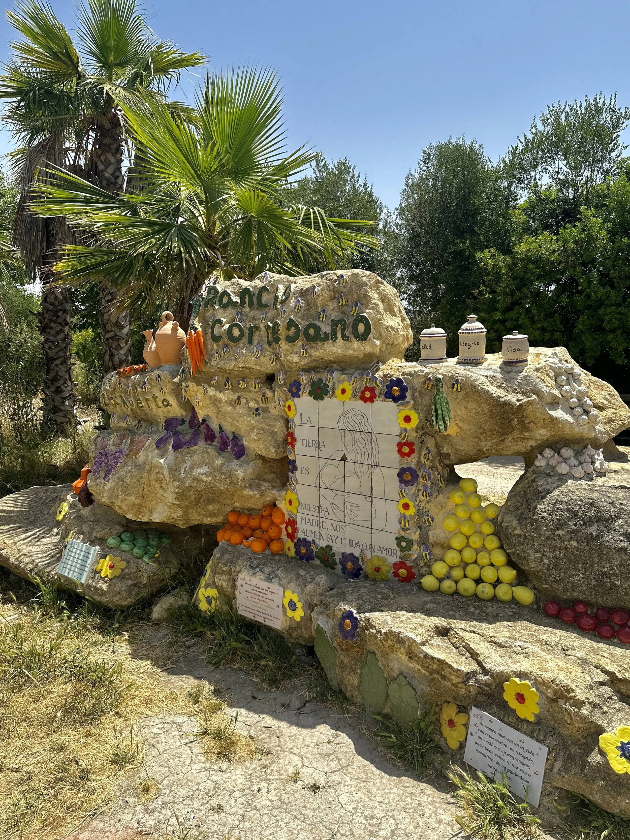 A memorial site with rocks decorated with colorful flowers, candles, and ceramic jars, surrounded by trees and palm plants in a sunny outdoor setting.