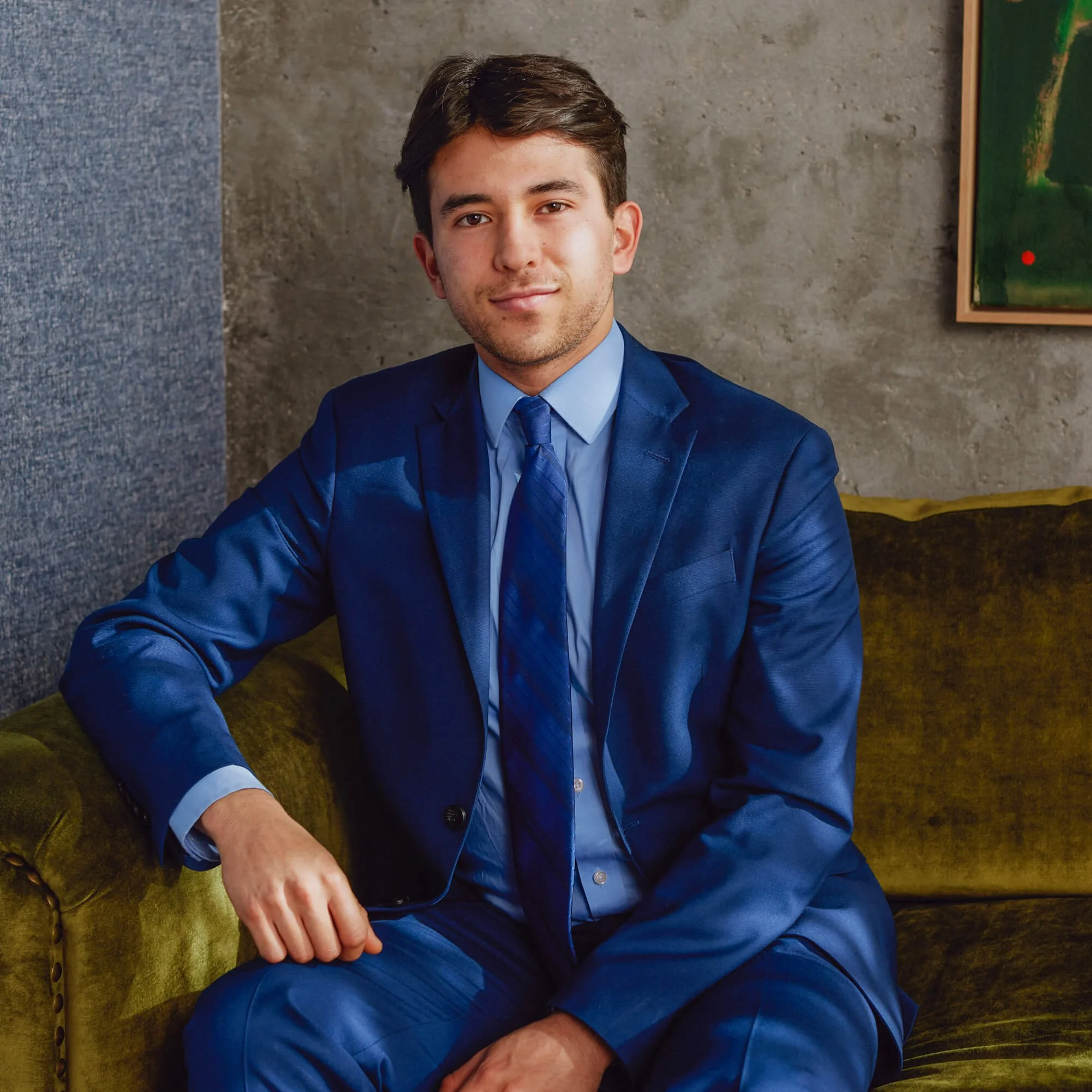 Juan Vargas, a young marketing specialist and analytics consultant with brown hair, wearing a blue suit and matching tie, sitting on a mustard-colored velvet sofa in a room with a gray concrete wall and colorful abstract paintings.