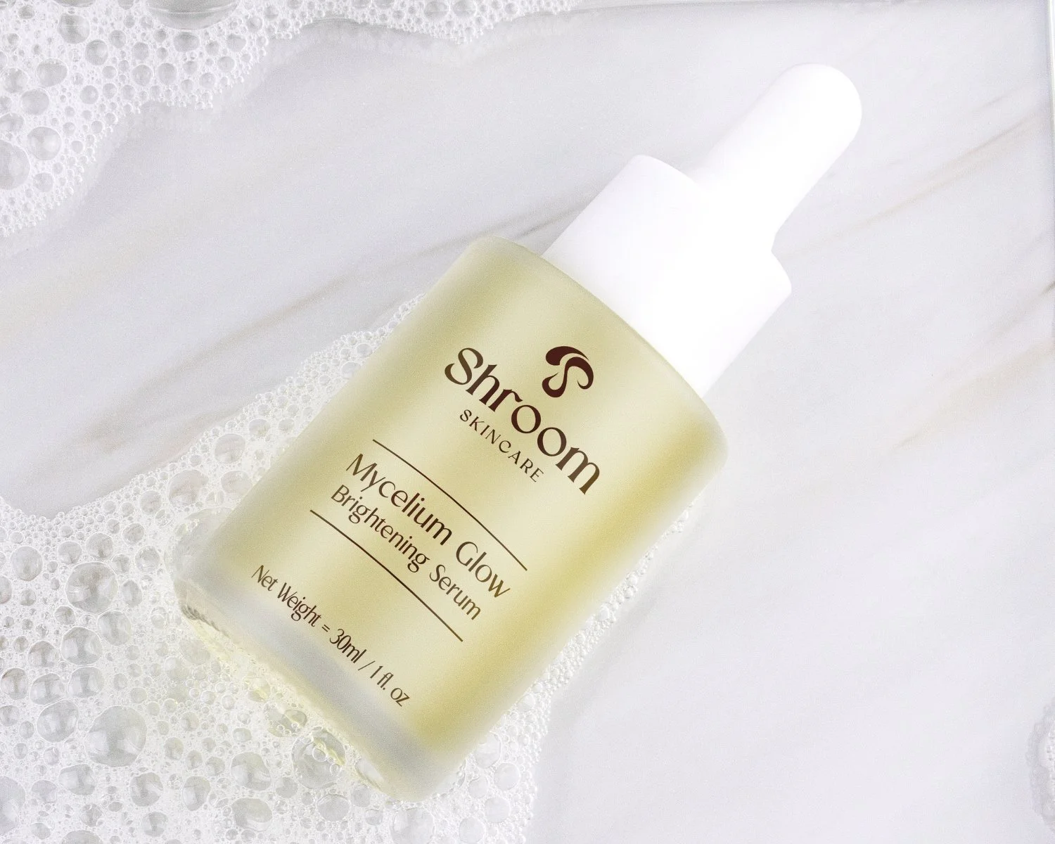A bottle of Shroom Skincare Mycelium Glow Brightening Serum placed on a white surface with soapy foam around it.
