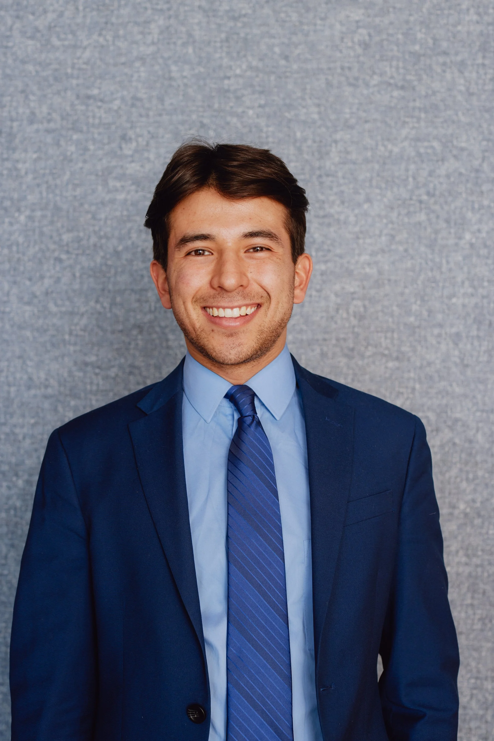 A young man with dark hair, light skin, and a friendly smile, dressed in a dark blue suit, light blue shirt, and a blue striped tie, standing in front of a gray textured background.