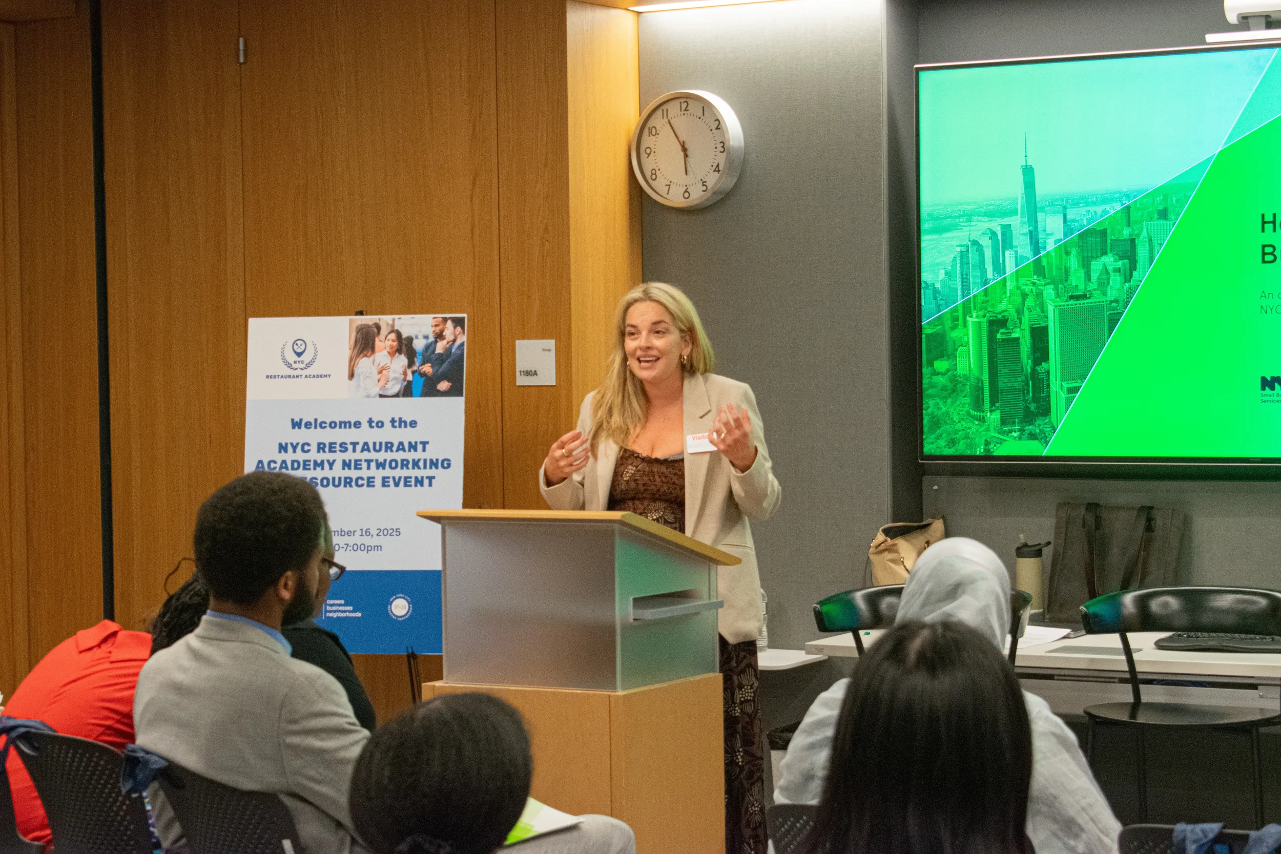 A woman with blonde hair and a beige blazer is speaking at a podium during a conference or seminar. There is a large digital screen beside her displaying a cityscape image. In the background, a sign indicates the event is organized by the NYC Restaurant Academy Network. Attendees are seated, listening to her, and a clock on the wall shows the time is approximately 10:09.
