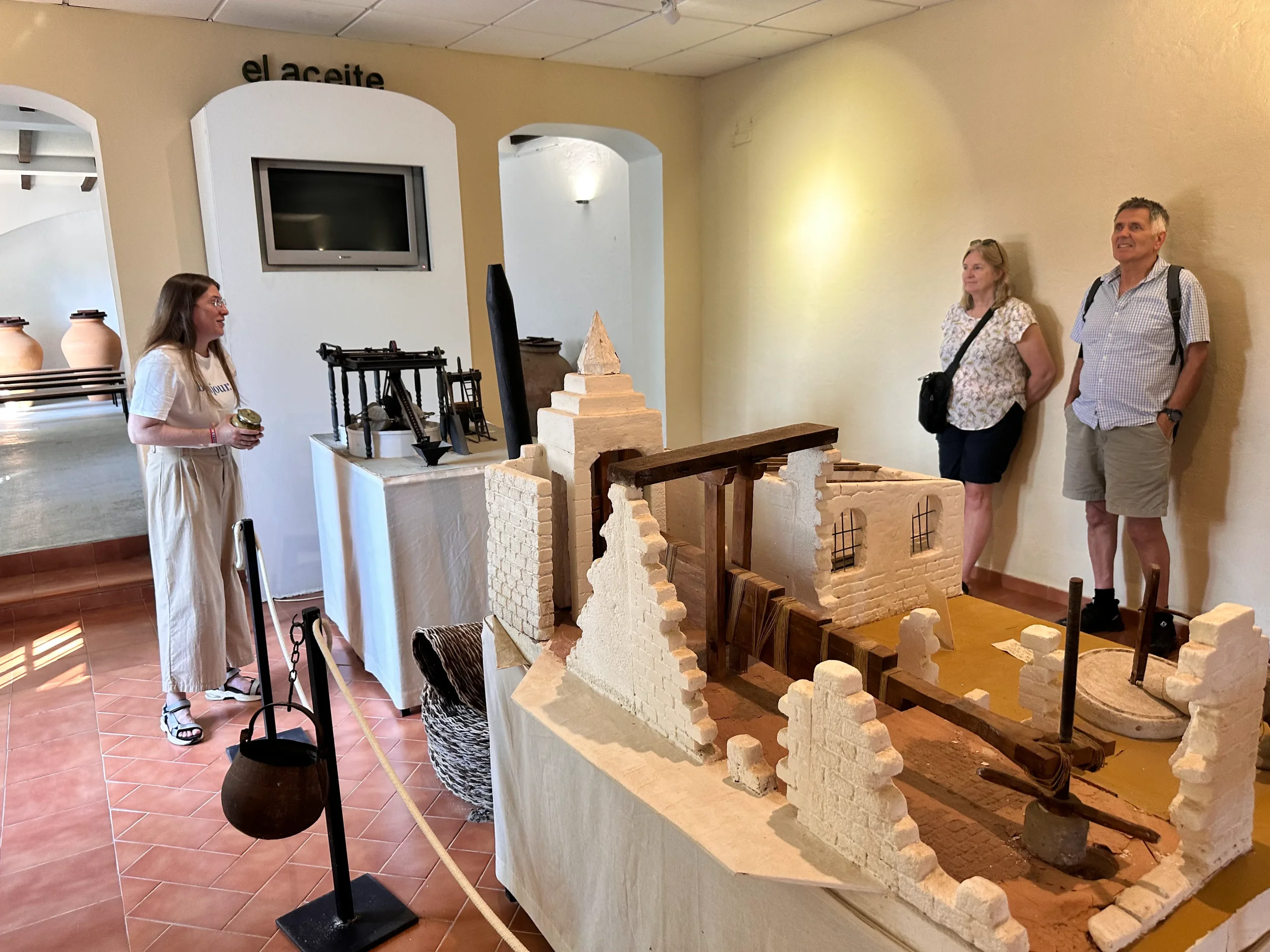 Three people in a museum observe a detailed model of ancient or colonial architecture made from stone and wood. The model features walls, arches, and small towers. There are display signs and a TV screen on the wall behind them.