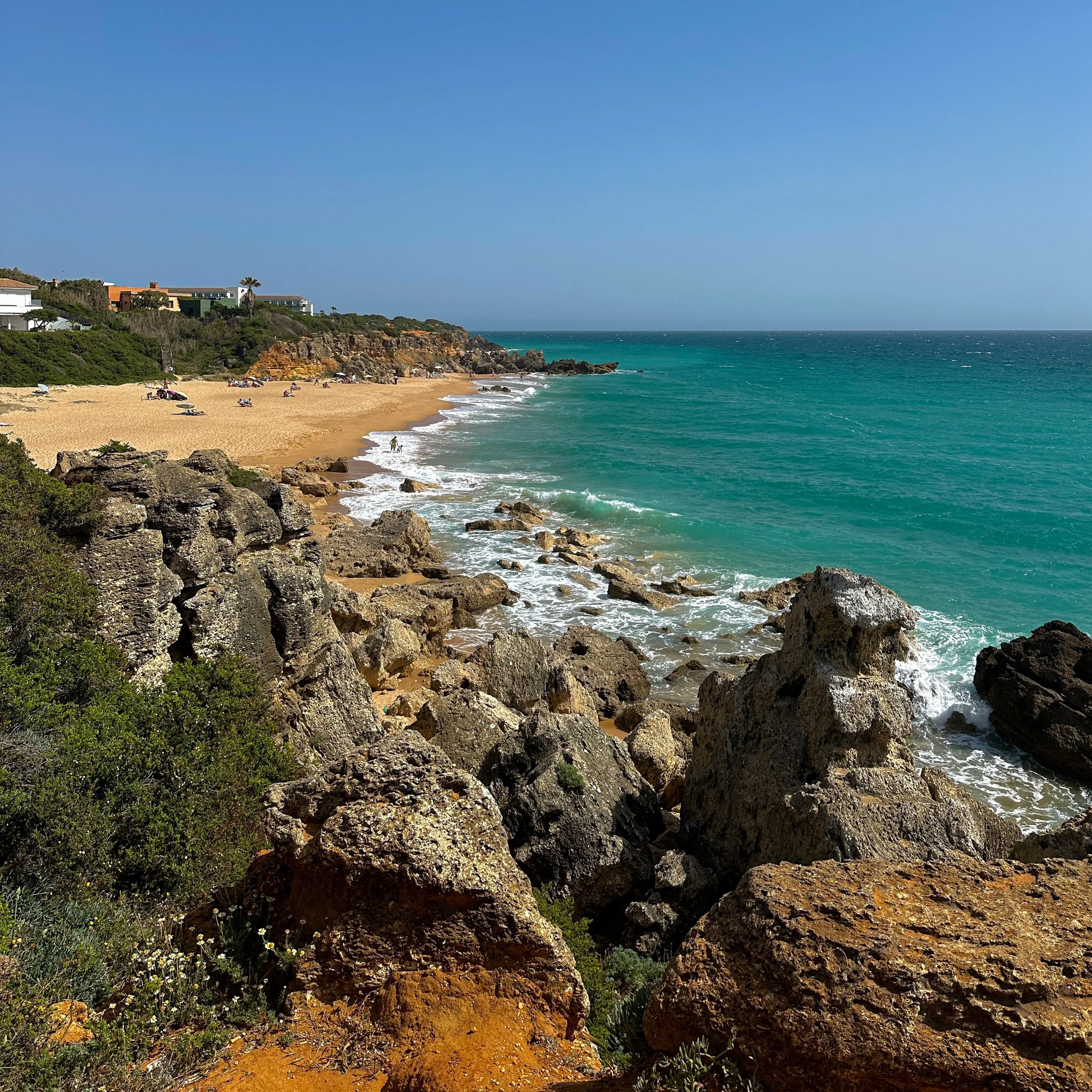 Scenic coastal landscape featuring rocky cliffs, sandy beach with sunbathers, clear turquoise water, and residential houses on the hillside under a blue sky.