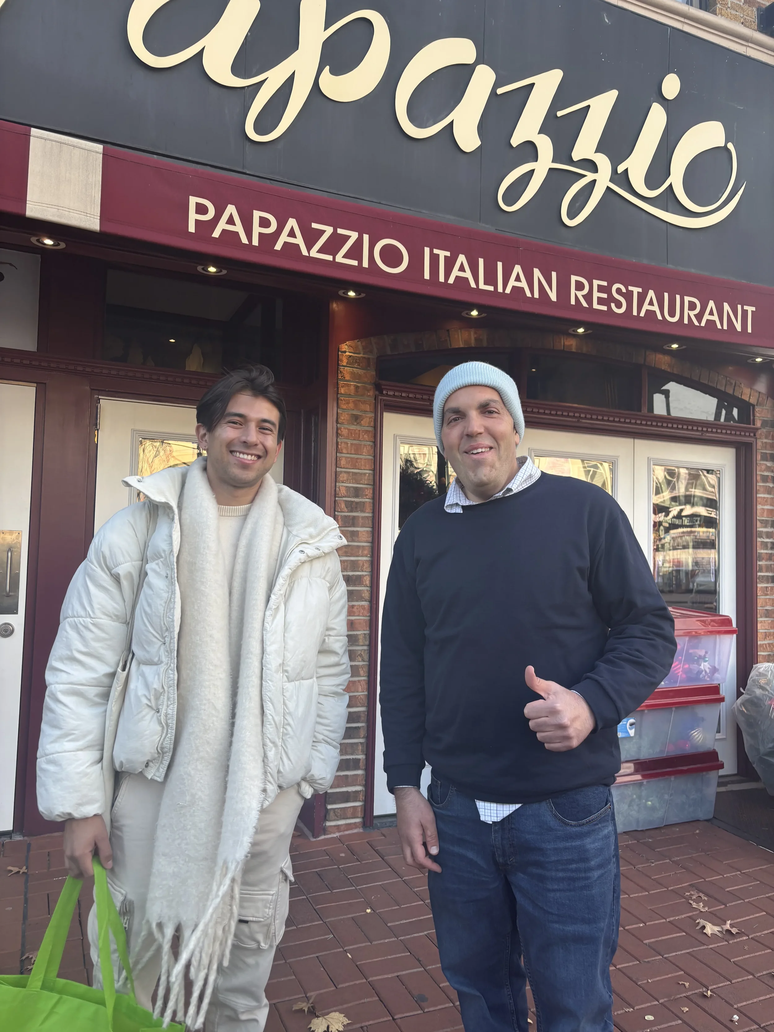 Two men standing in front of an Italian restaurant called 'PAPAZZIO'. One man is wearing a white puffy coat and scarf, and the other is wearing a black sweater, plaid shirt, and a beanie. They are smiling and posing for the photo.