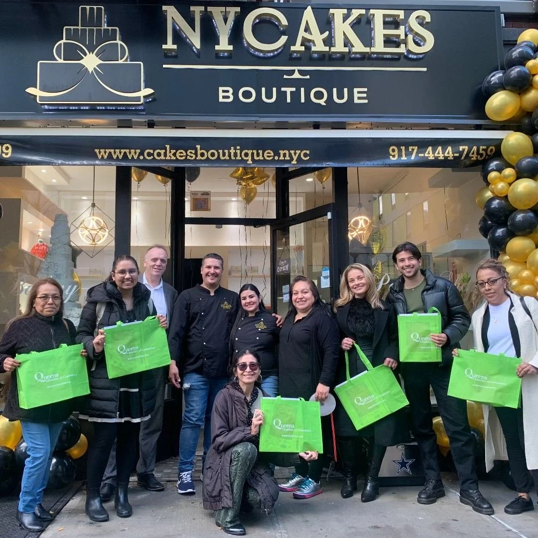 Group of ten people standing and kneeling outside a cake boutique, holding green shopping bags with the logo 'Queens.' The storefront has a black and gold sign reading 'NYCAKES BOUTIQUE' and a decorative cake logo, with gold and black balloon decorations on the right.