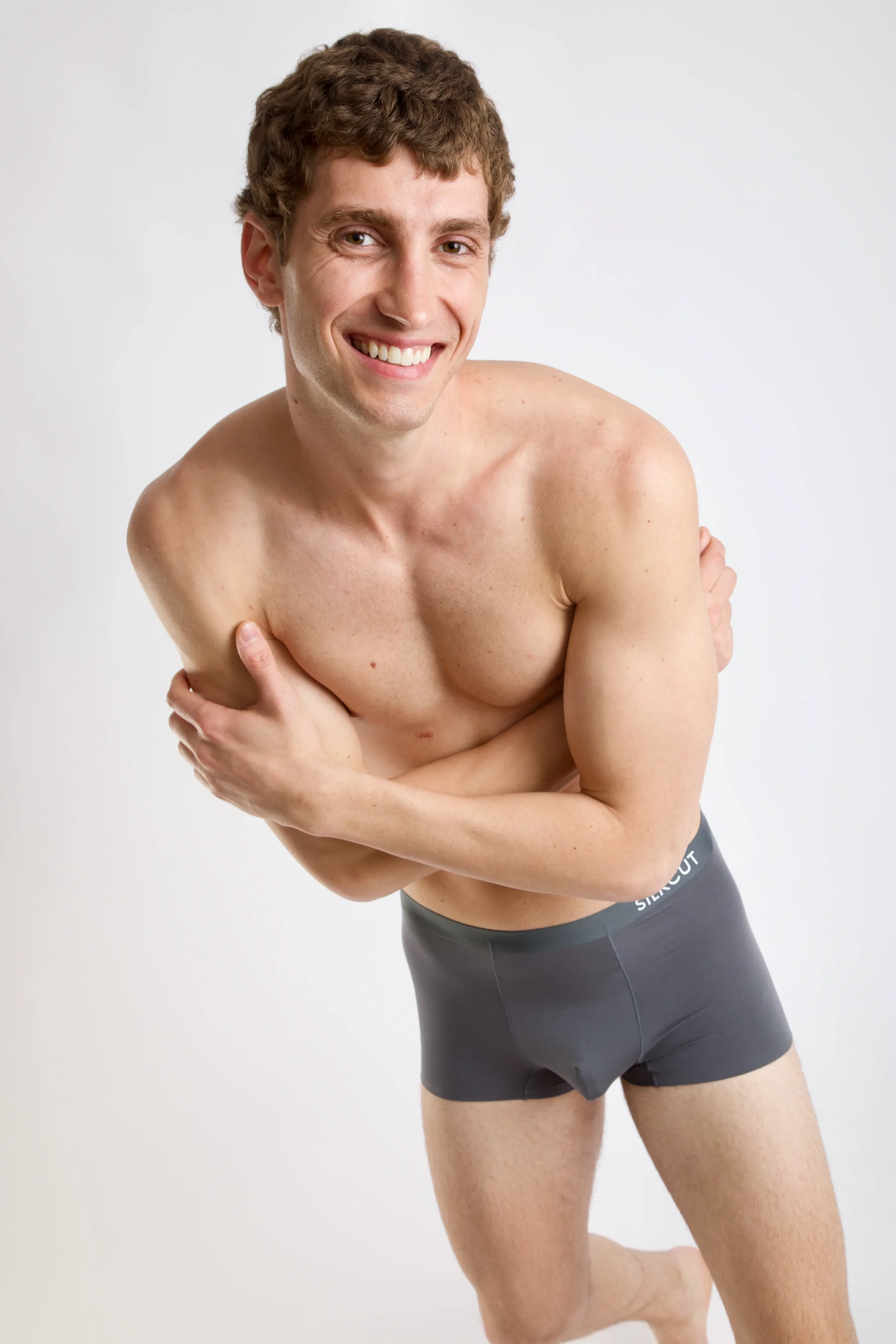 Mid-shot of a smiling male model with crossed arms wearing grey Silkcut boxer briefs in a white-backdrop studio shoot.