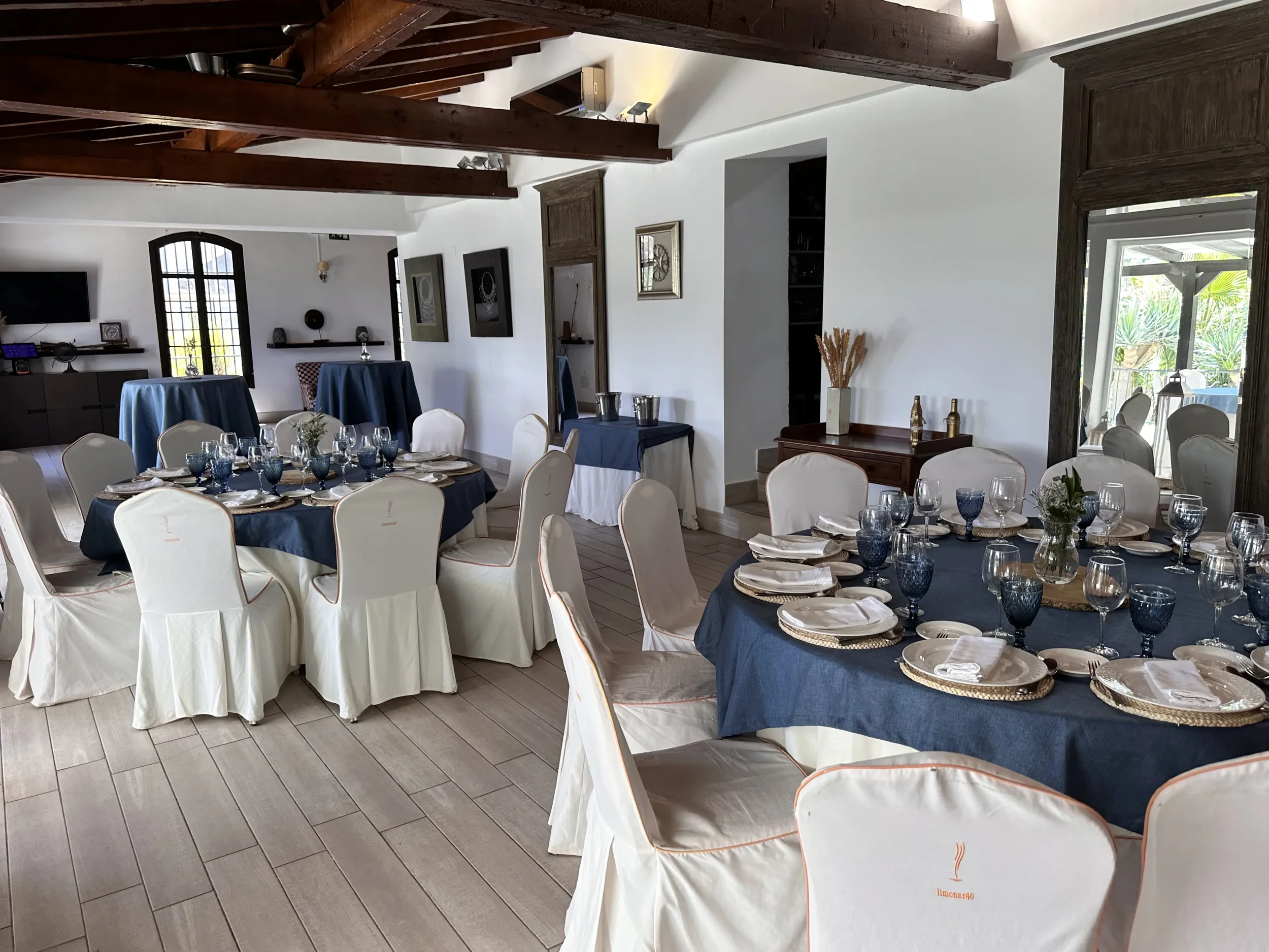 A dining room set up for a celebration with tables covered in blue tablecloths, white chairs with beige slipcovers, and tables set with blue glasses, white plates, and silverware. The room has white walls, dark wood beams, artwork on the walls, and a door leading to a patio with greenery outside.