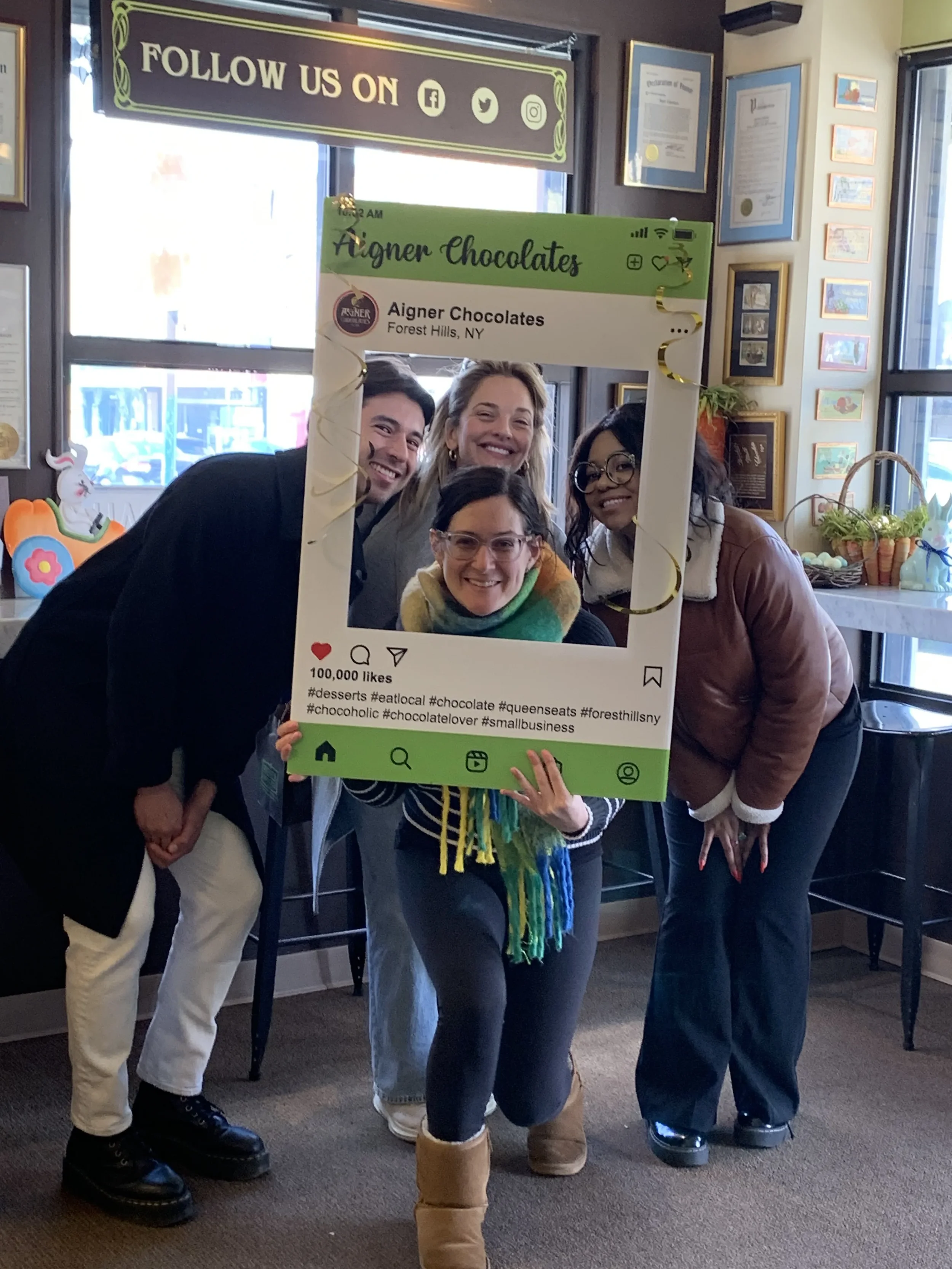 Four women posing together behind a Instagram-style photo frame at a chocolate shop, with colorful decorations and certificates on the wall.
