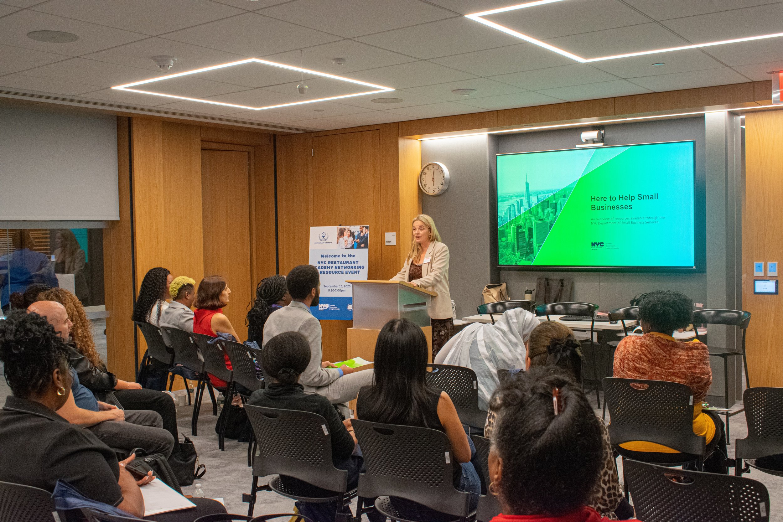 A woman speaker presenting at a conference with a large screen displaying a green-themed presentation slide that reads "Here to Help Small Businesses". Audience members are sitting and listening in a modern conference room.