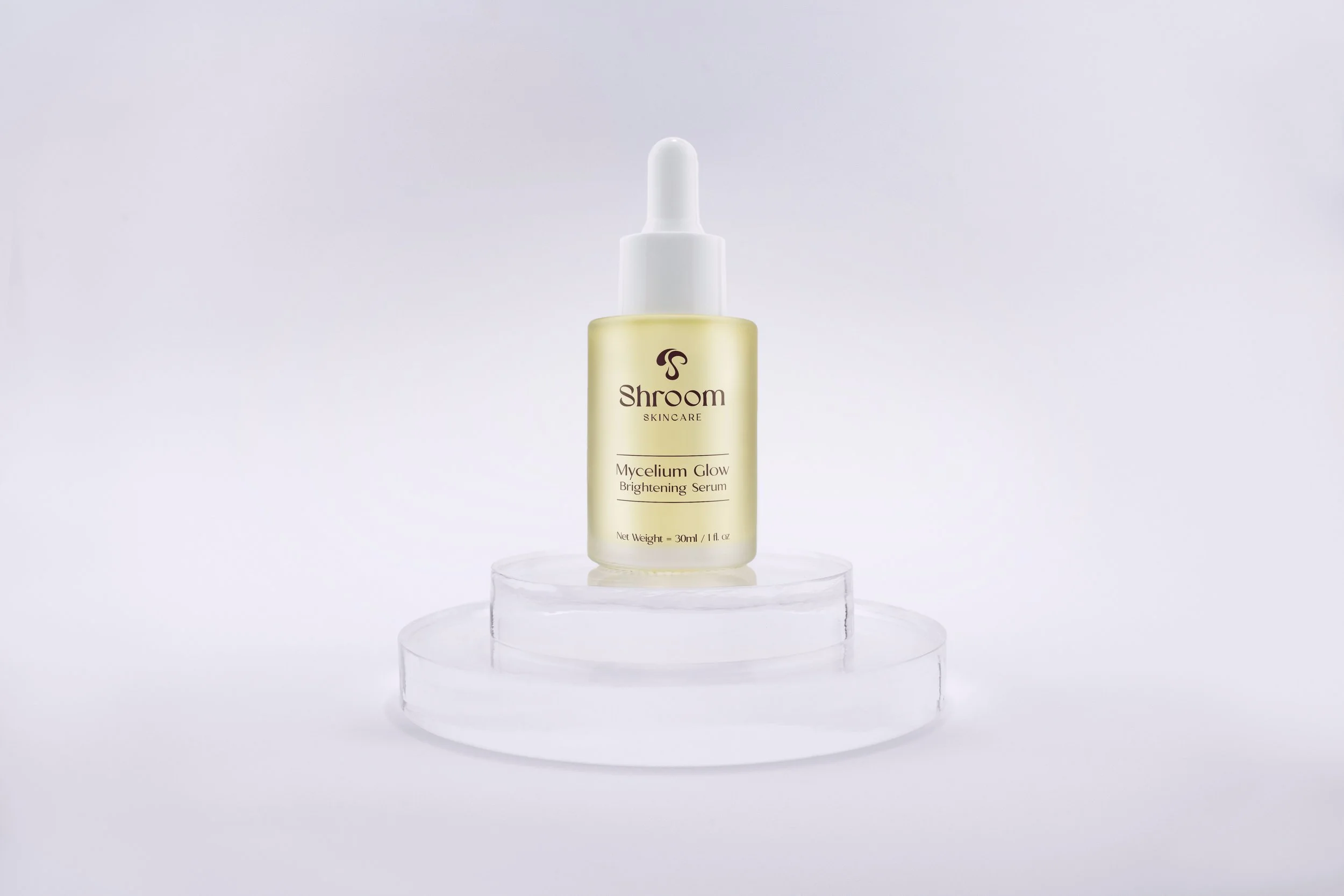 A small bottle of Sroom Skincare Mycelium Glow Brightening Serum on a clear, round platform against a white background.