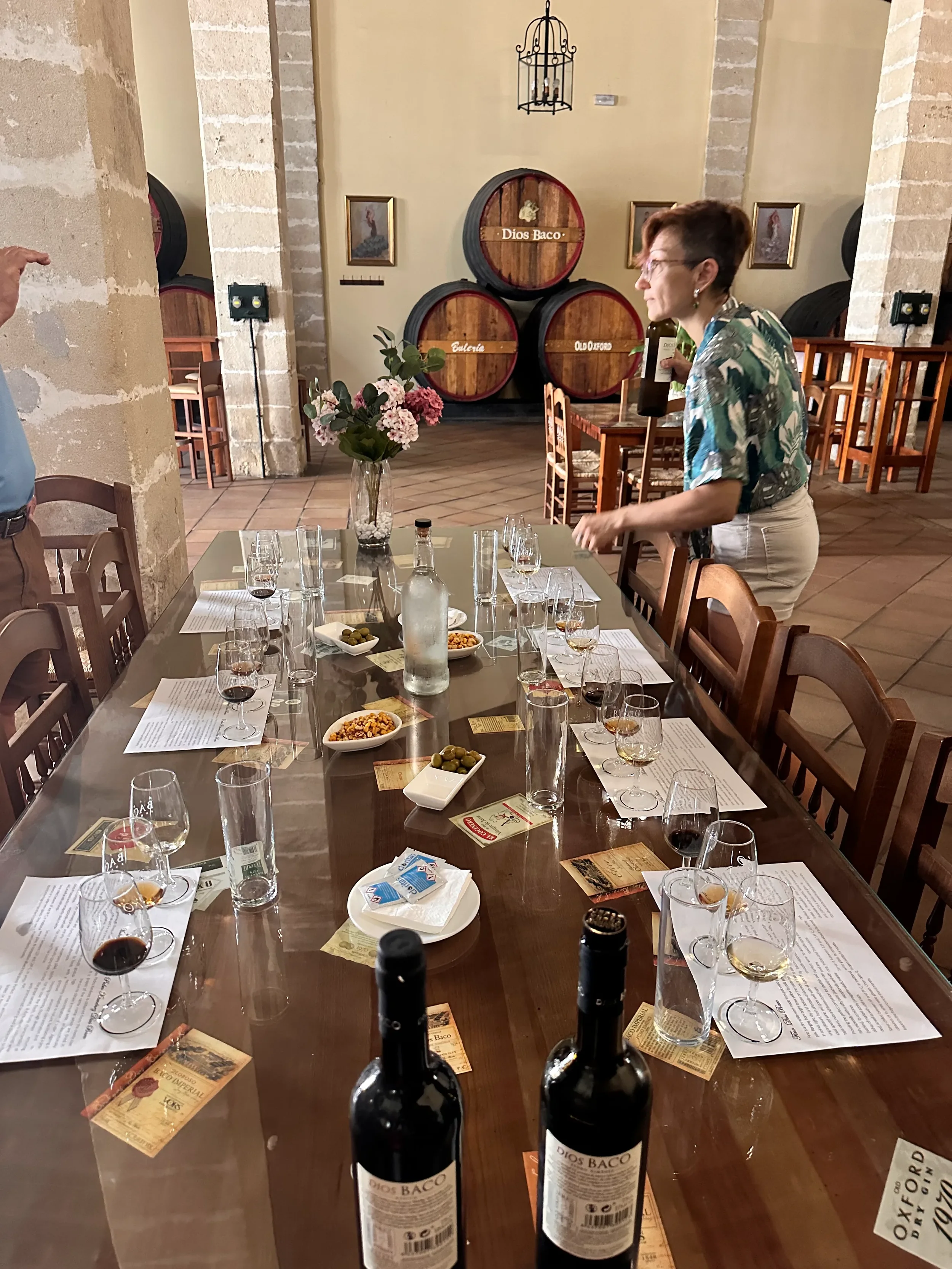 A dining table set for a wine tasting with multiple wine glasses, bottles of wine, bowls of snacks, and informational sheets. In the background, a woman is holding a bottle of wine, with wine barrels labeled 'Dios Baco,' 'Eulalia,' and 'Old Oxford' visible. The room features exposed brick walls and framed artwork.