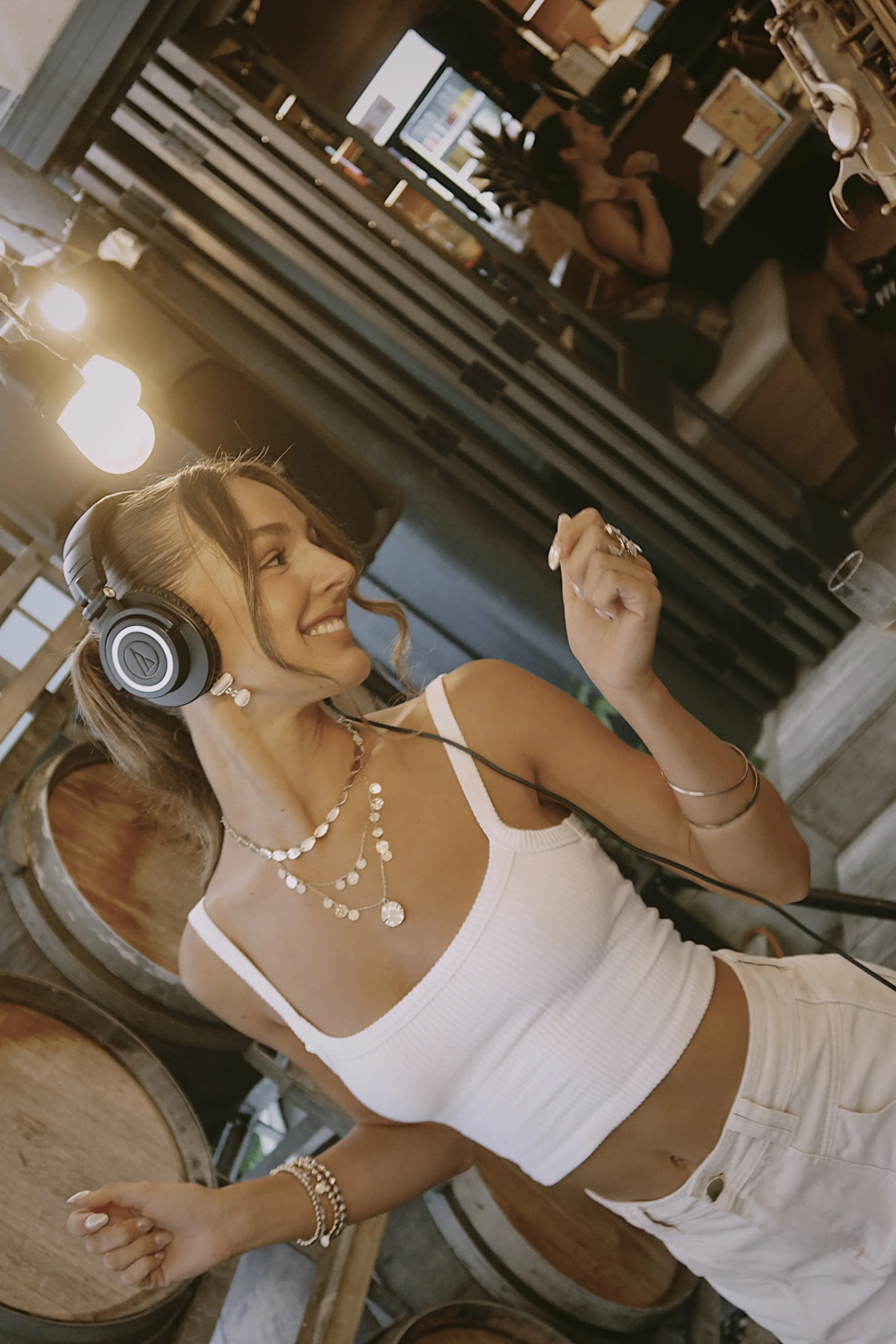 A woman with headphones, wearing a white tank top and beige pants, dancing and smiling in a cozy, dimly lit room with a rustic decor.