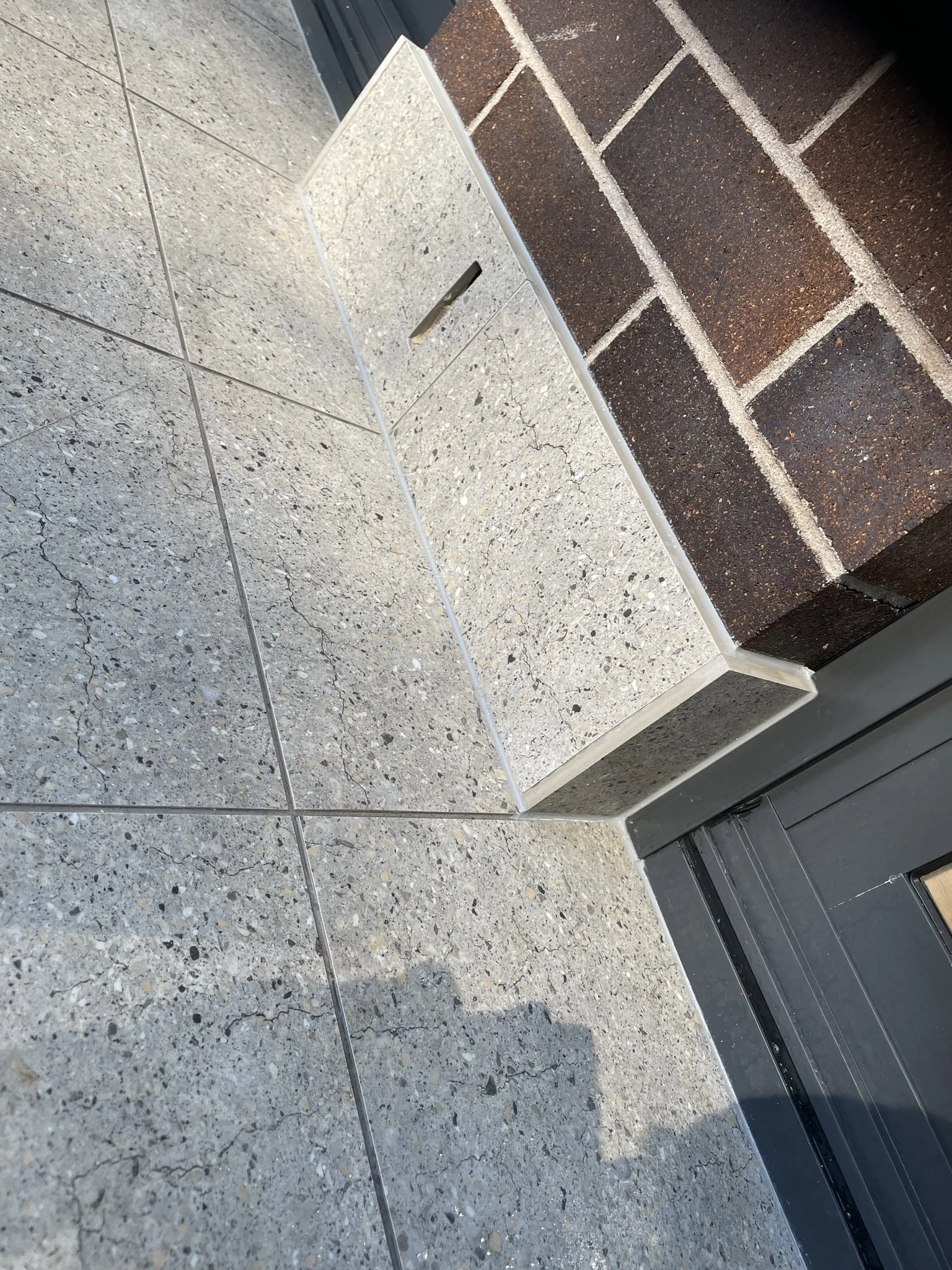 Close-up of tiled floor and wall with a small floor vent near a brick wall and a dark-colored door frame.