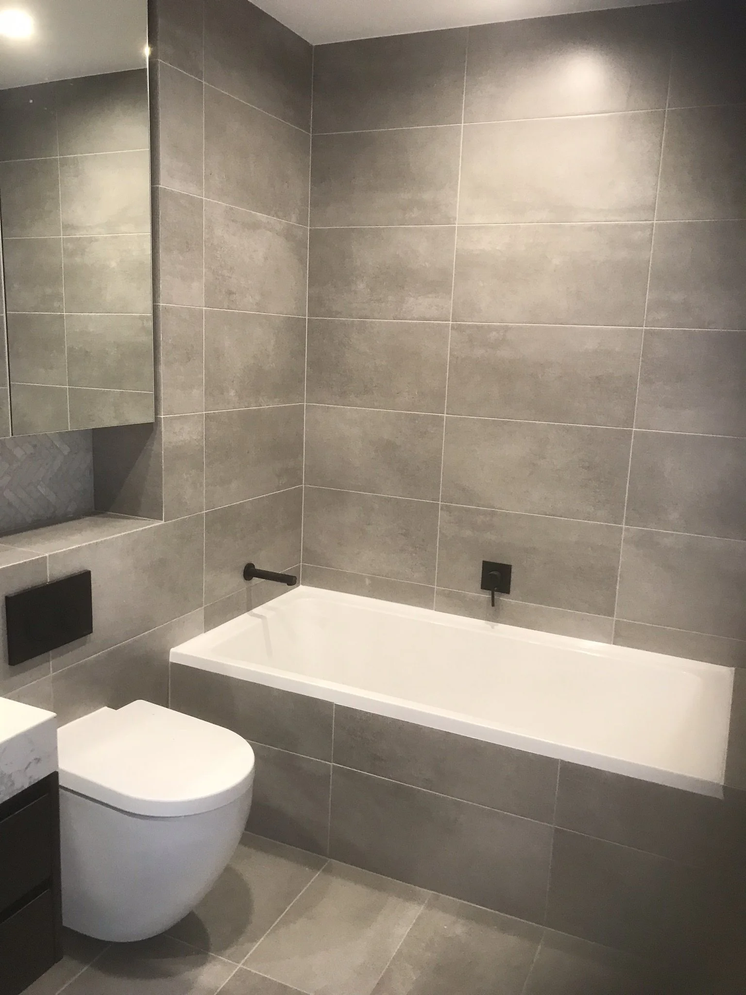 Modern bathroom with gray tiles, a white bathtub, a wall-mounted black toilet, and a mirror.