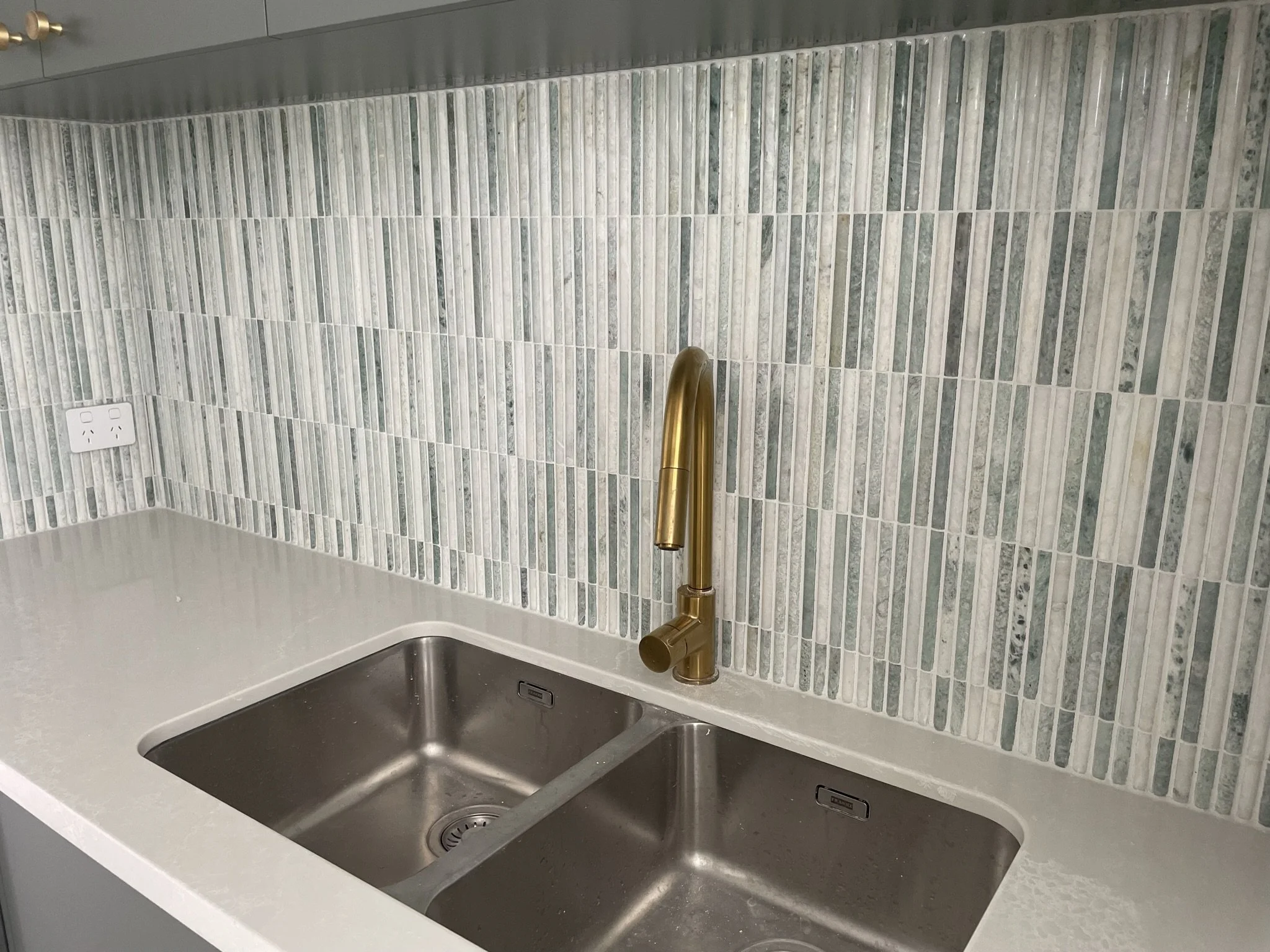 Kitchen sink with two basins, a gold-colored faucet, a light-colored countertop, and a mosaic tile backsplash in shades of green and gray. An electrical outlet is visible in the corner.