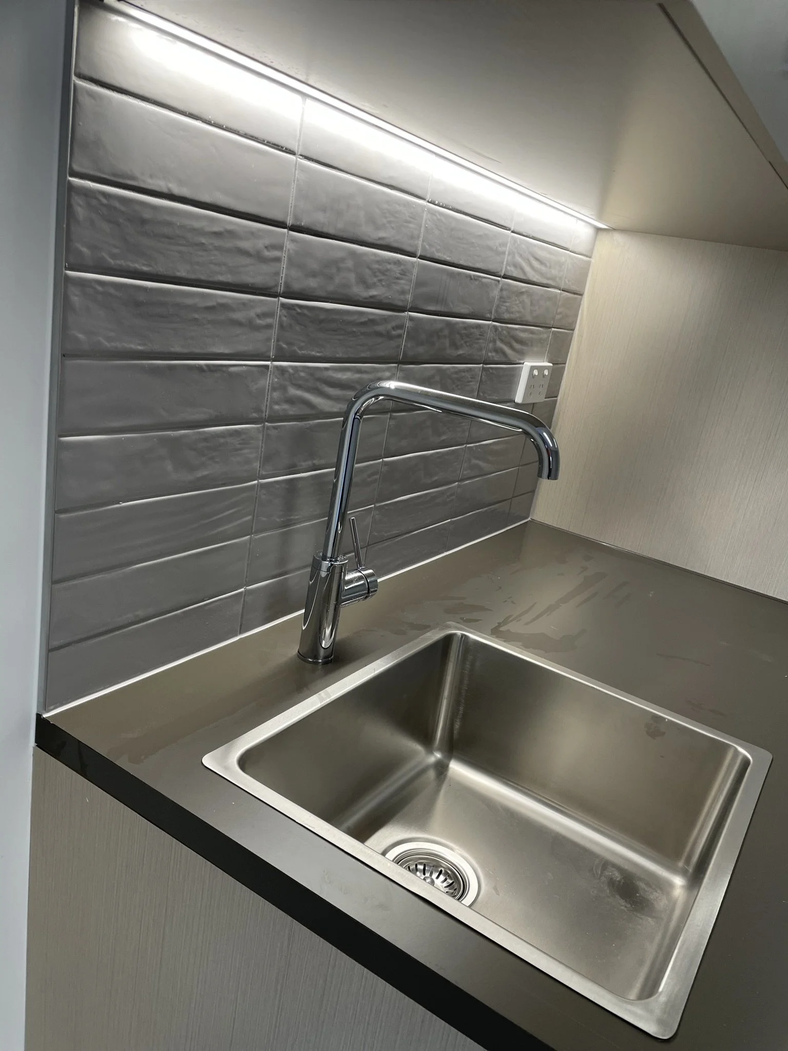 A stainless steel kitchen sink with a tall, curved faucet, set in a beige countertop with a metallic backsplash, and a power outlet on the wall.
