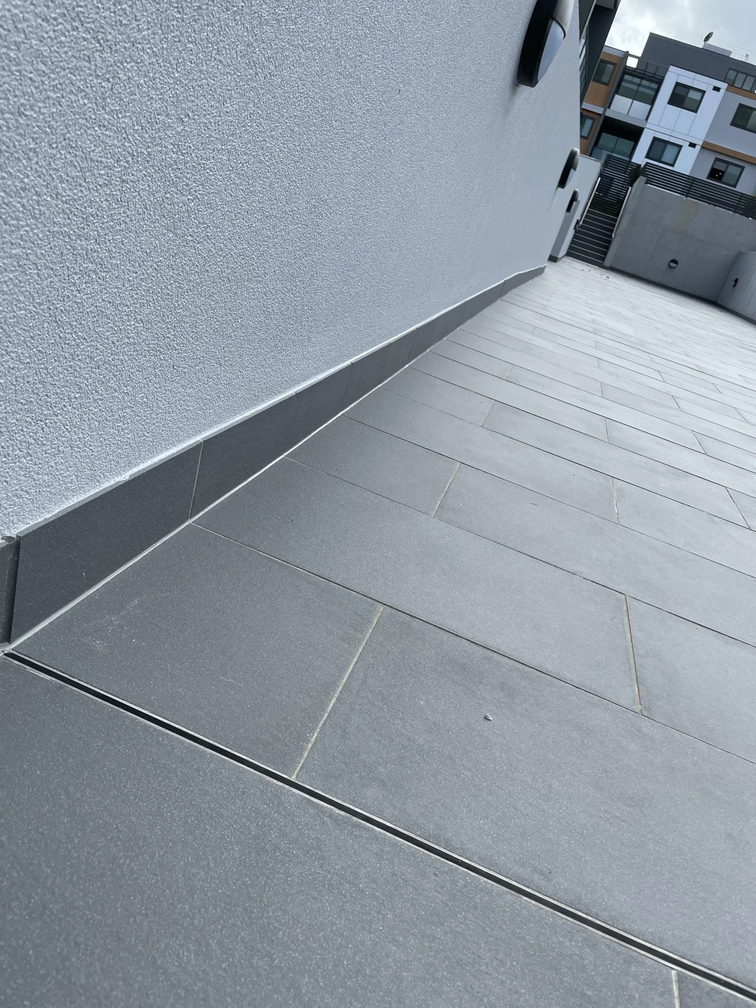 Exterior view of a modern building with textured white wall and gray tiled ground, featuring a drain trap along the base of the wall and a small black object on the ground.