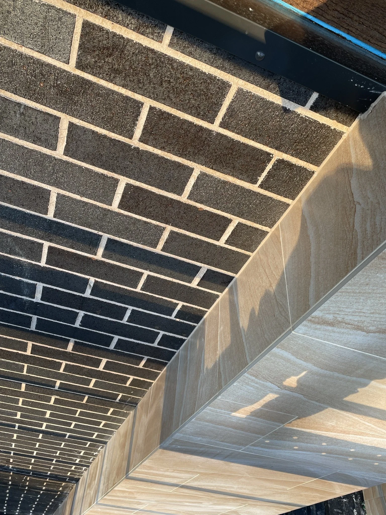 Close-up view of a corner showing a brick wall with dark bricks and light mortar, next to a tiled wall and floor with tan and beige tiles.