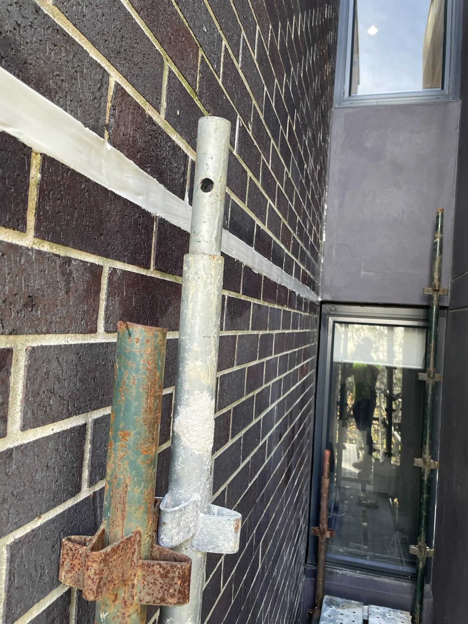 Close-up of scaffolding pipes mounted on a dark brick wall, with a window and reflection of a person visible in the background.