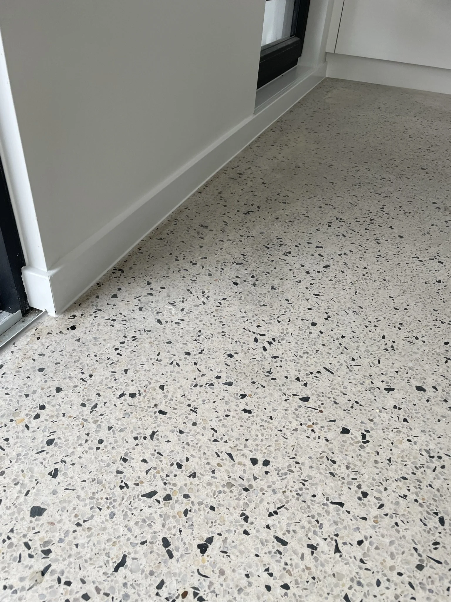 Close-up of a polished terrazzo floor near a door and wall, with a beige base and black, gray, and brown chips.