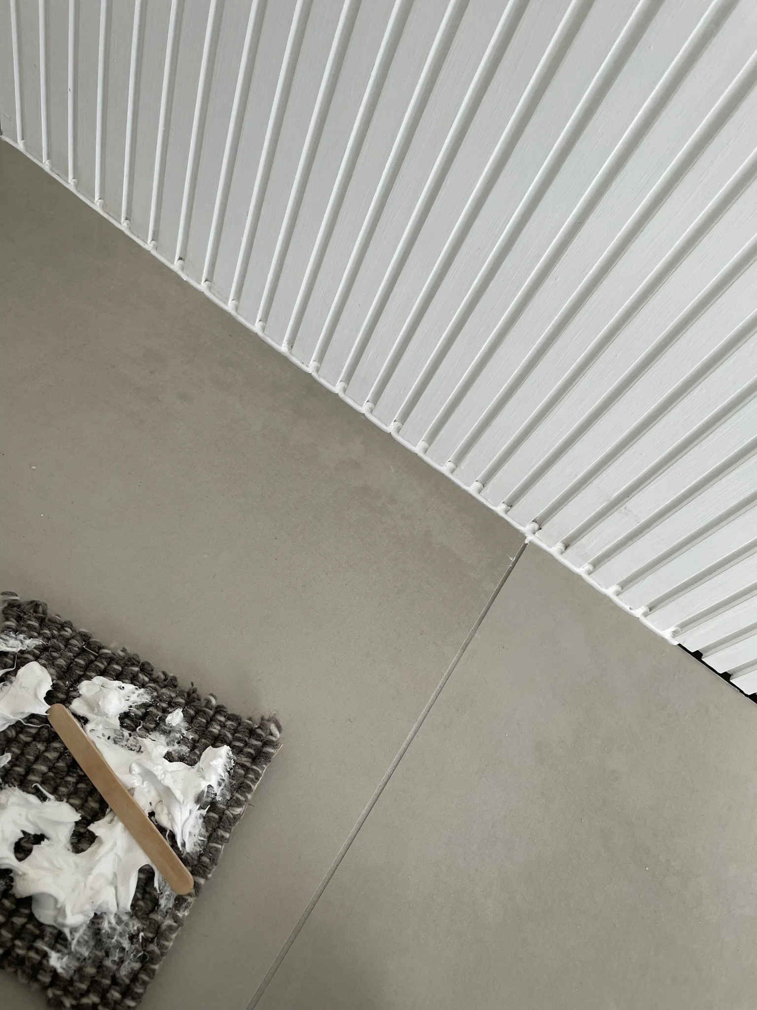 A corner of a room with a textured white ceiling with vertical grooves, a smooth grey wall, and a grey floor. There is a black and white textured mat with white foam and a wooden stick on it.